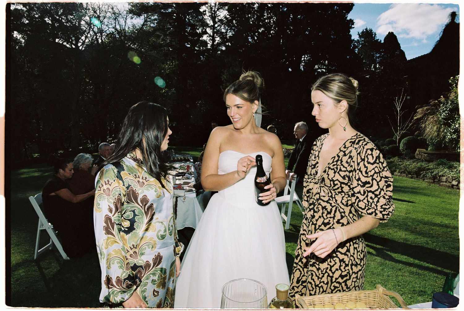 Bride in white gown chatting with two guests in floral outfits at an outdoor wedding reception.