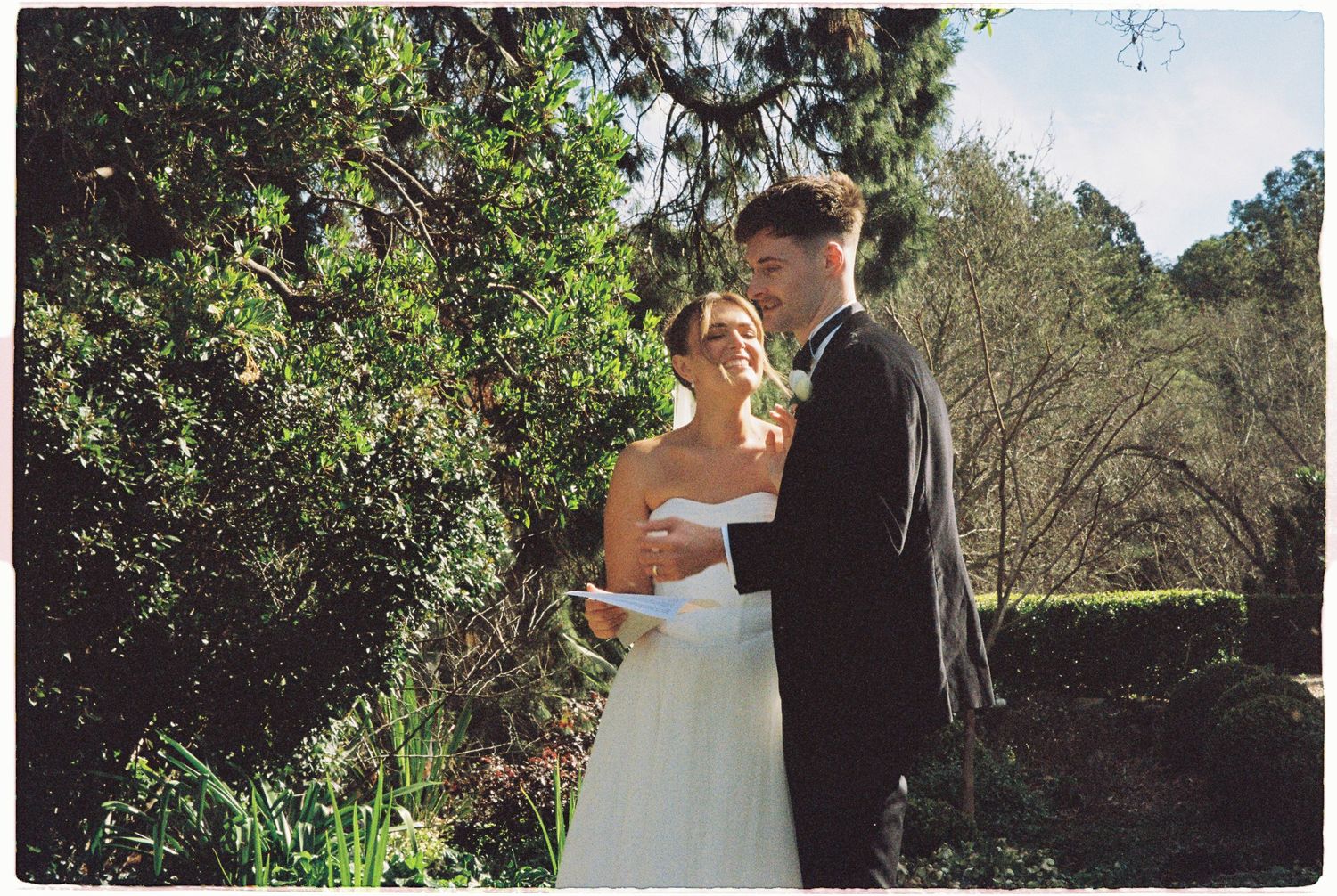 Bride and groom share tender moment in white and grey wedding attire surrounded by lush green garden trees.