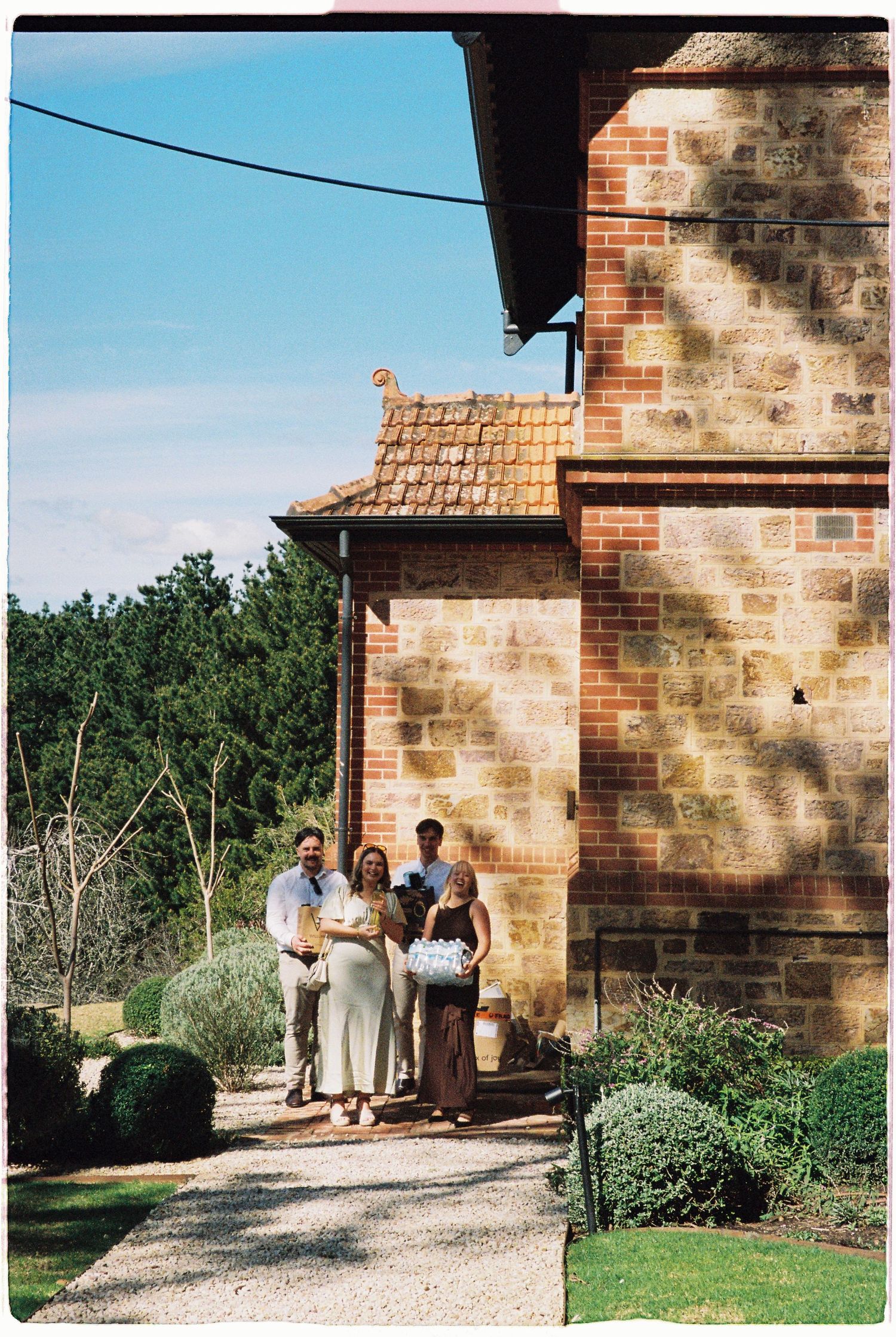 Family group posing outside a rustic stone brick building surrounded by lush gardens on a sunny day.