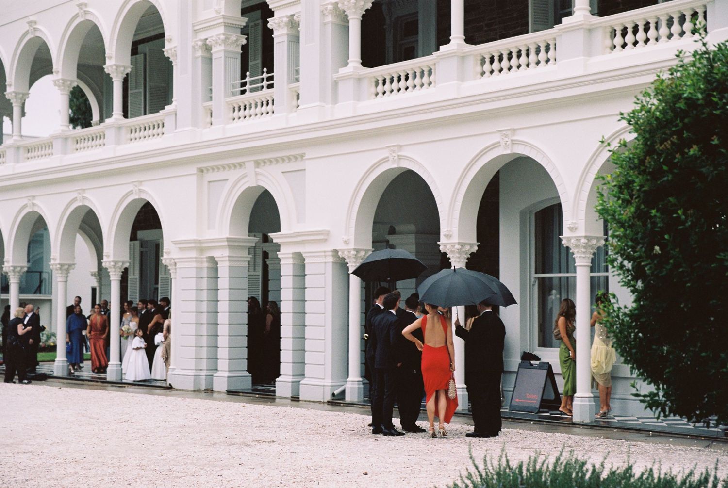 Wedding guests gathering under black umbrellas outside elegant white colonial building with arched colonnade.