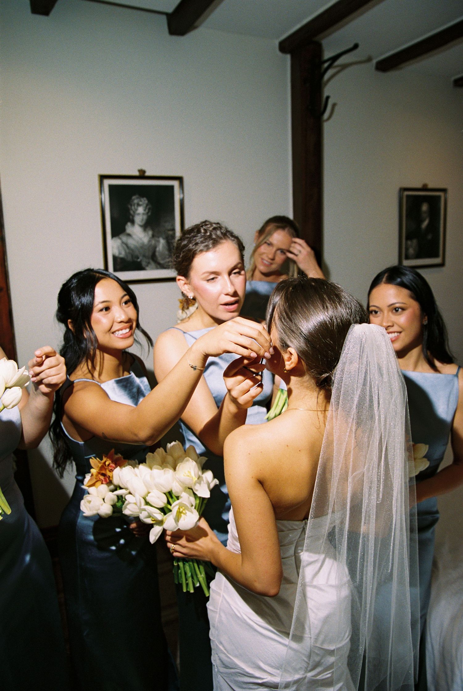Bridesmaids in blue satin dresses helping bride with makeup before wedding ceremony indoors.