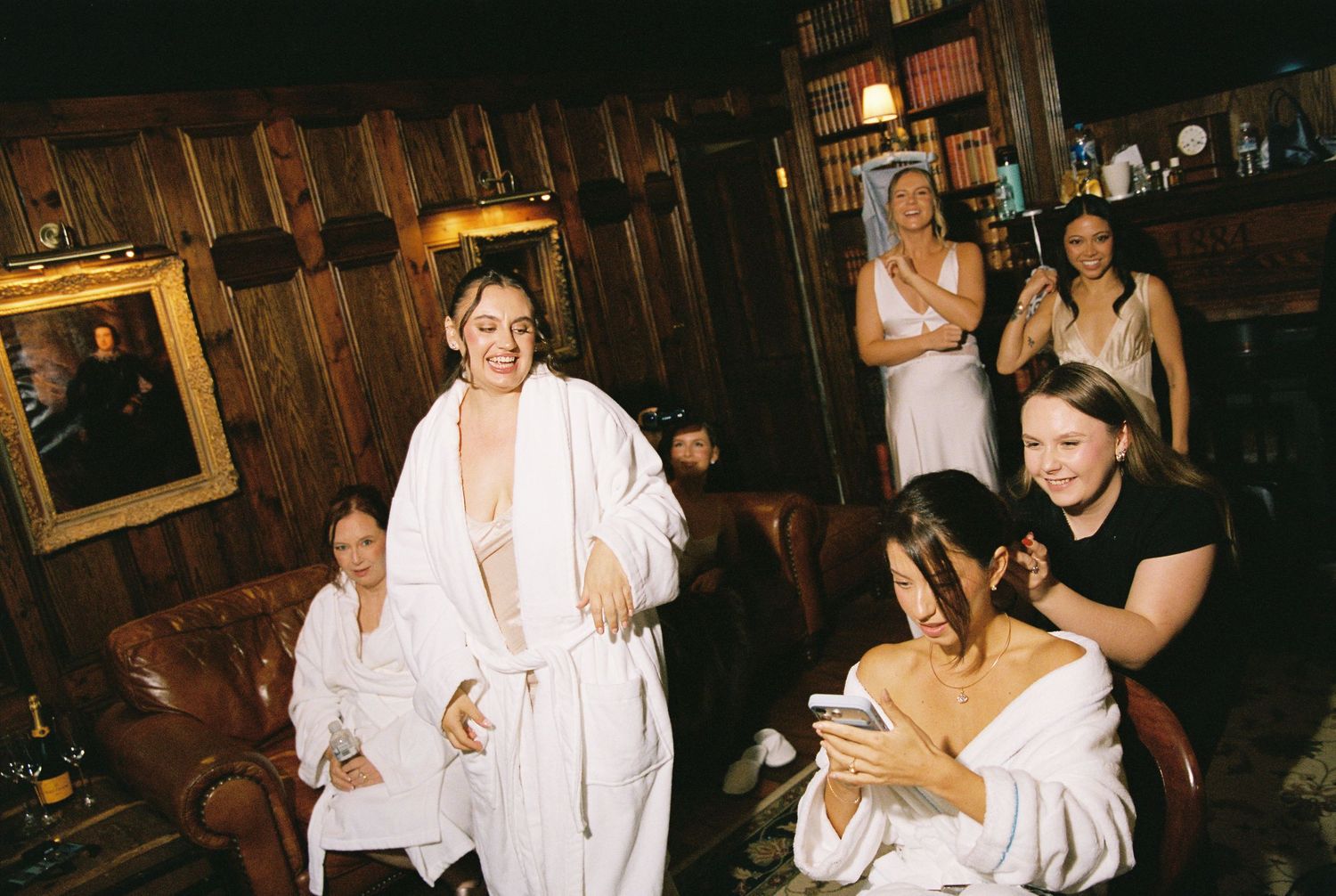 Bridal party in white robes laughing and getting ready in a dark wood-paneled venue.