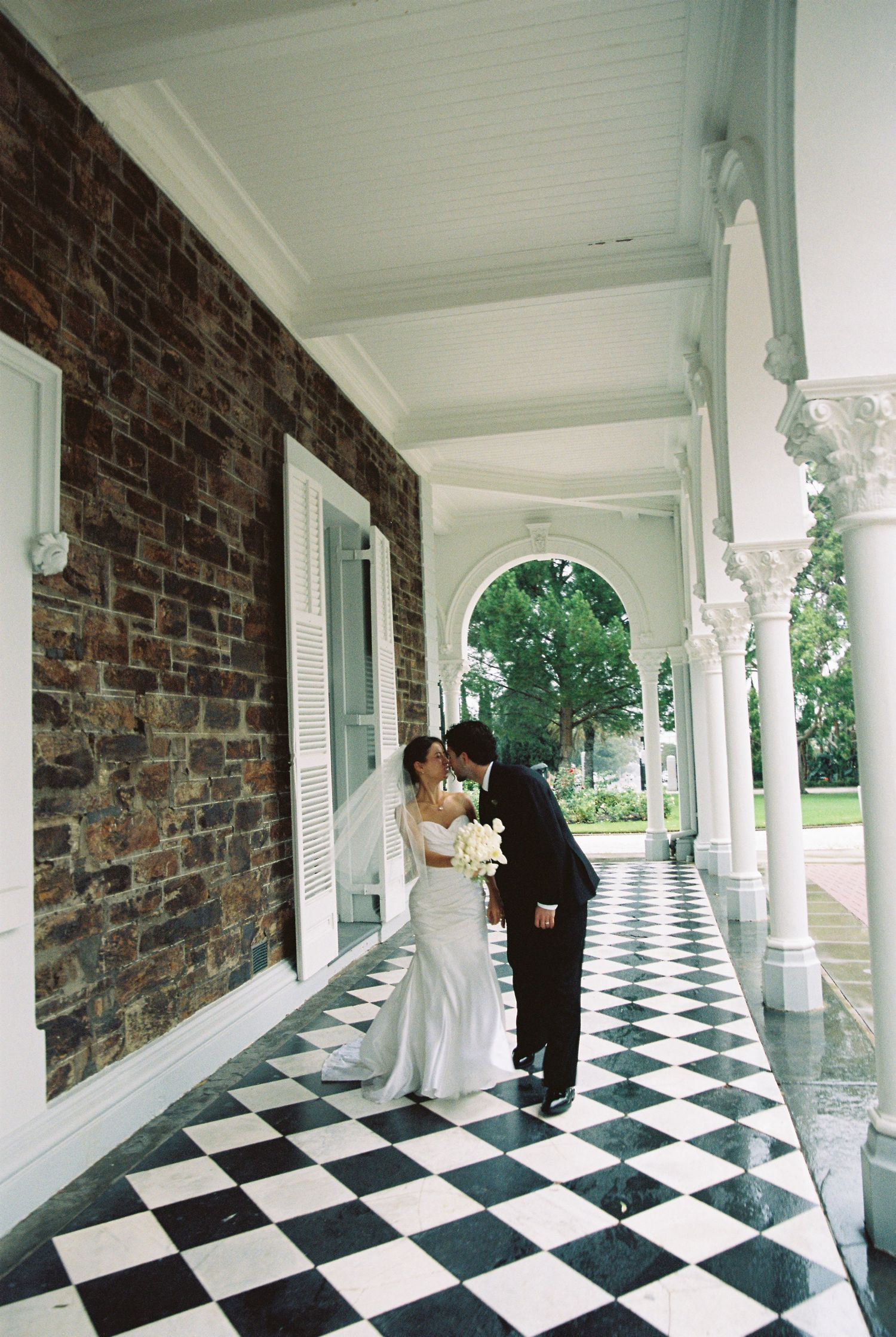 Bride and groom share a kiss on elegant columned porch with black and white checkered marble floor.