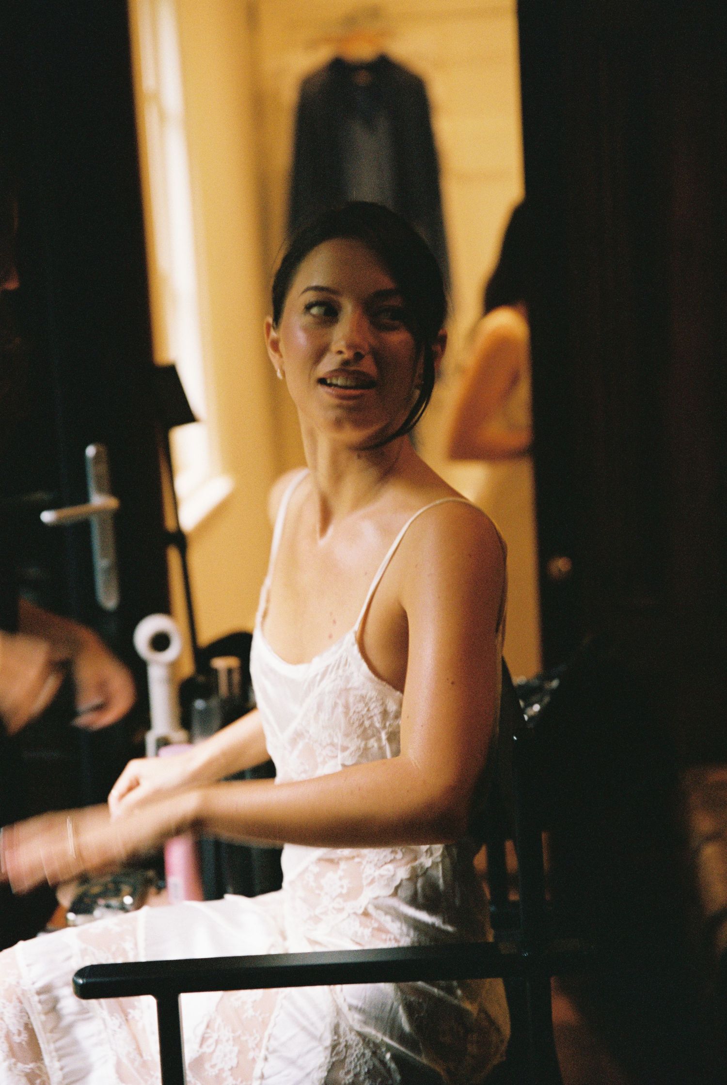 Beautiful bride in white lace slip dress sits relaxed during wedding preparations in a warmly lit room.