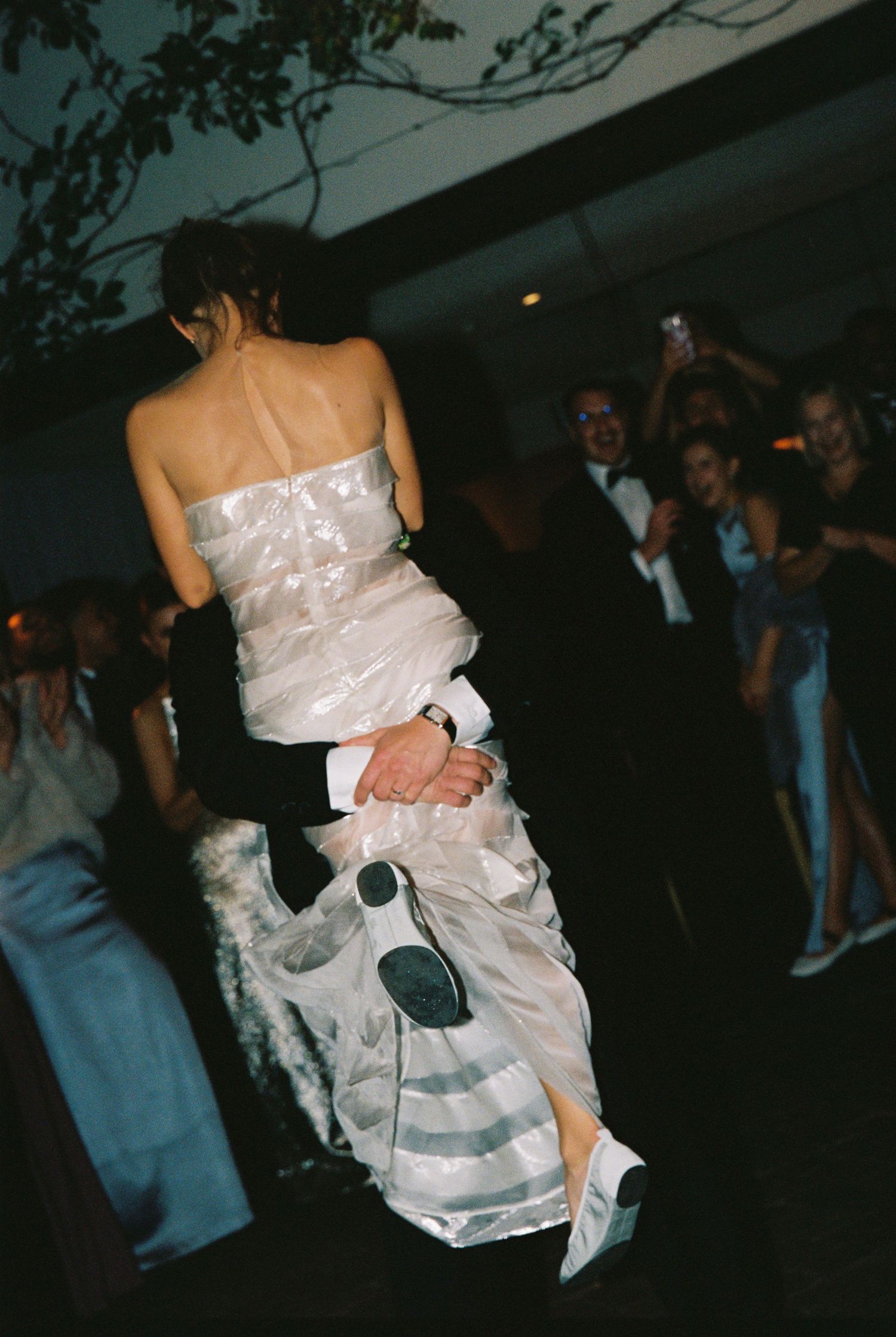 Groom lifting bride in wedding dress on dance floor surrounded by guests.