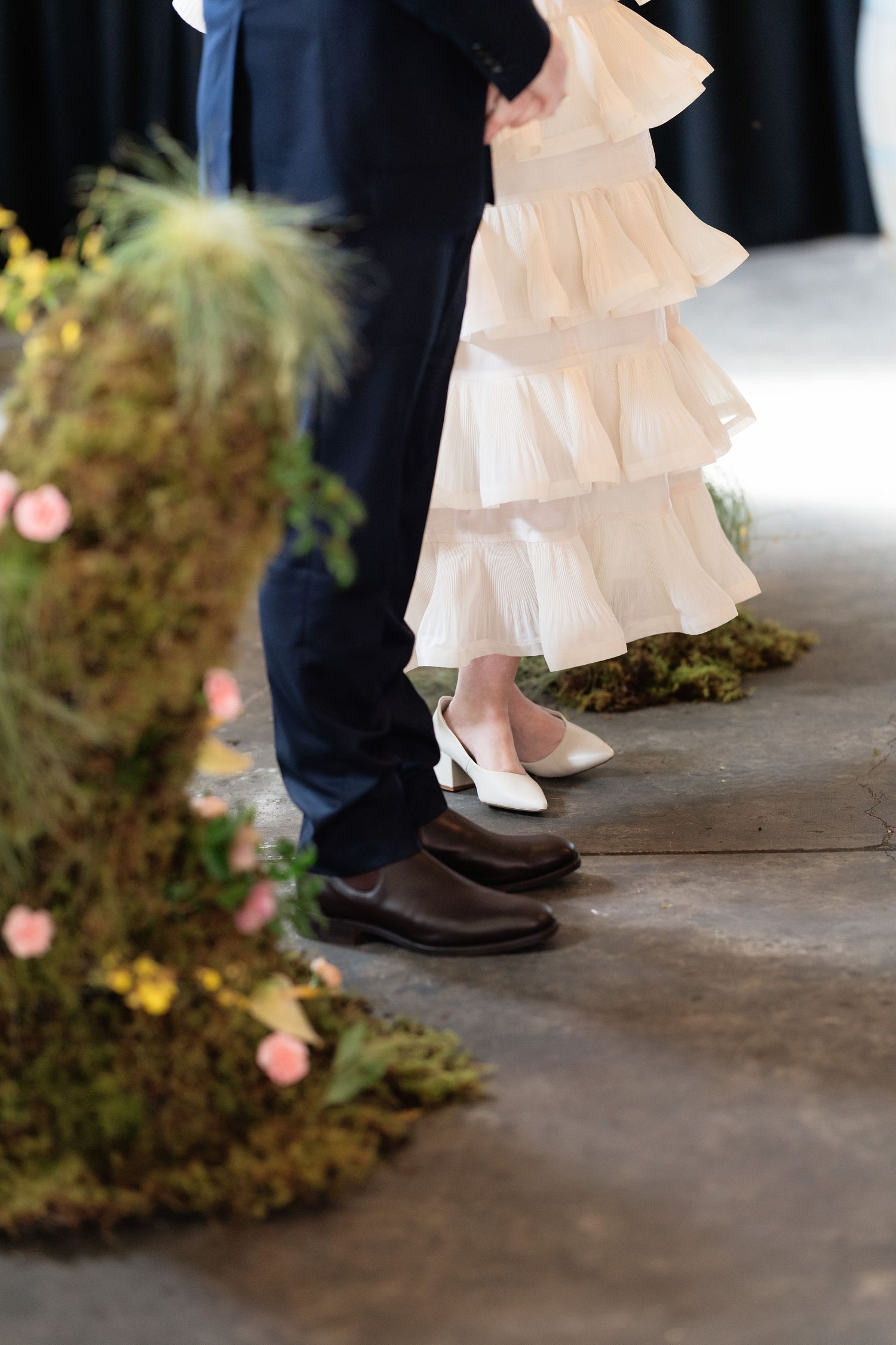Close-up of bride in ruffled white wedding dress and groom in navy suit standing by floral moss decor.