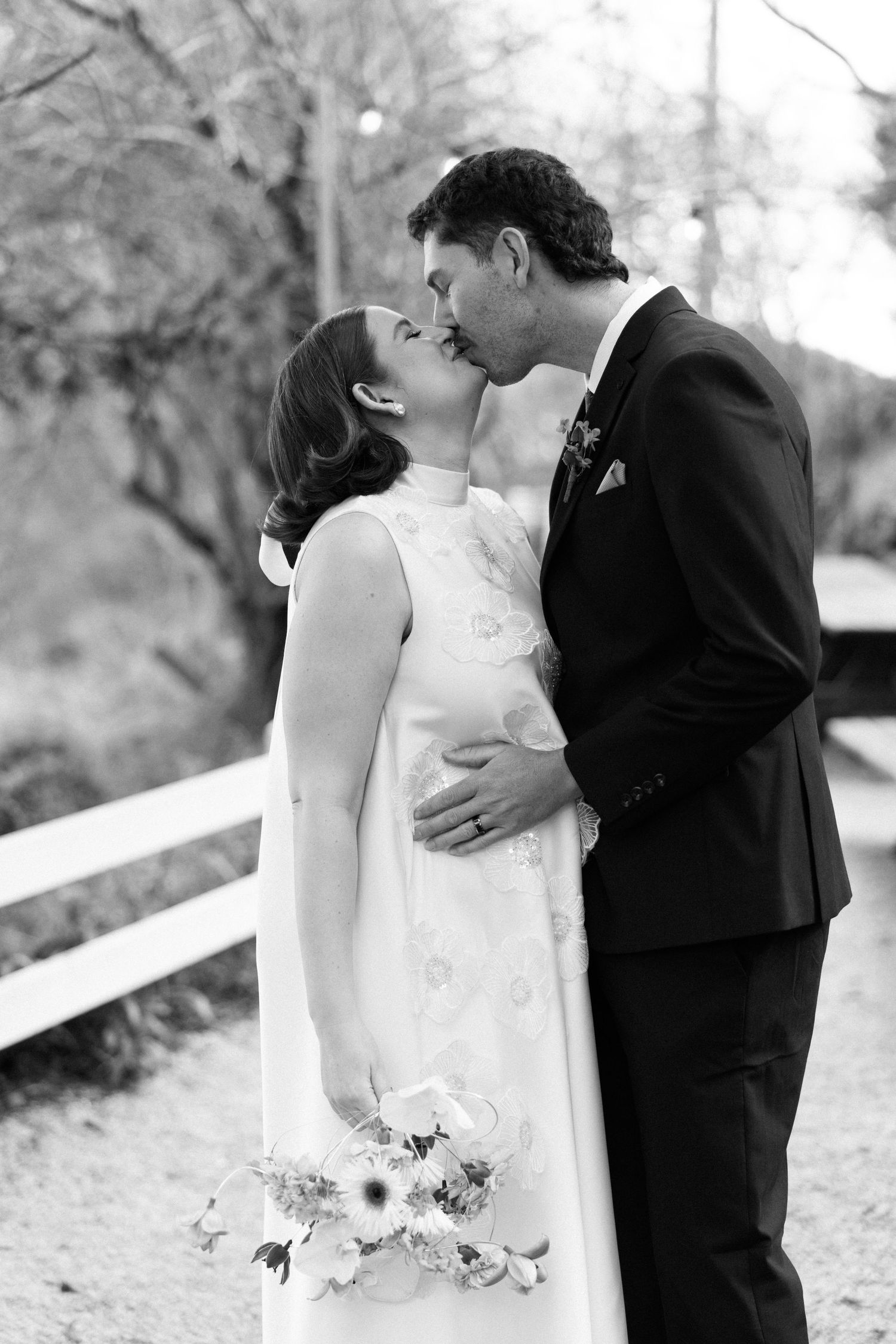 Black and white photo of a bride and groom sharing a kiss outdoors, bride holding a floral bouquet.