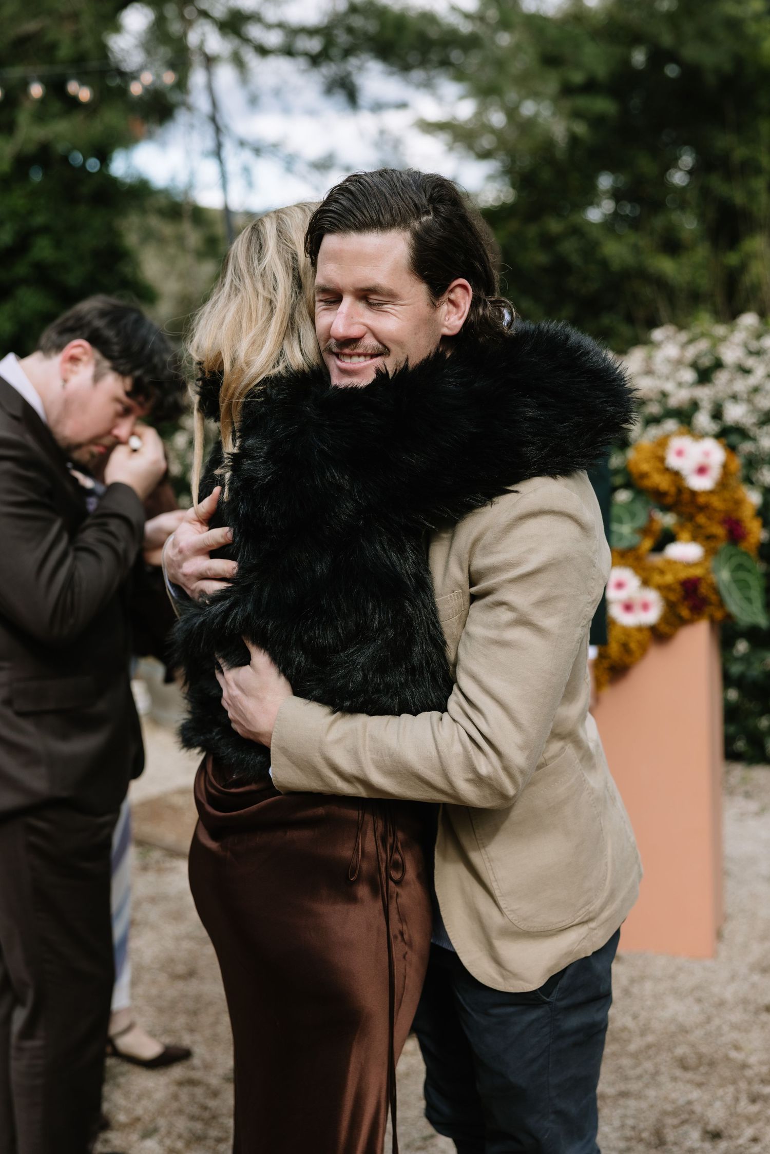A man in a tan jacket smiles while hugging a woman in a black fur vest at an outdoor wedding reception.