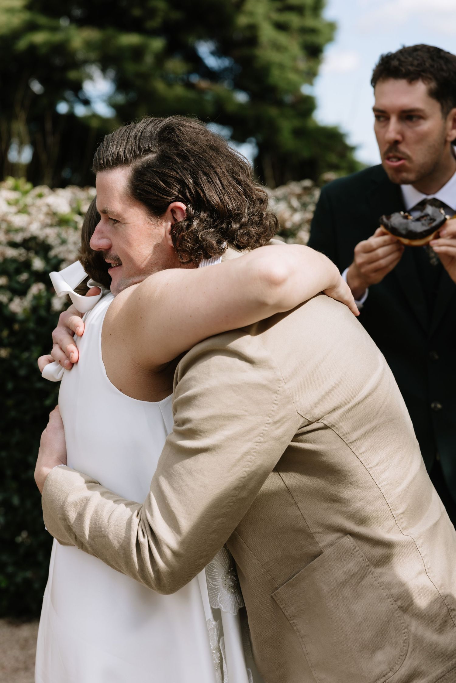 Emotional wedding hug between bride in white dress and guest in beige suit outdoors.