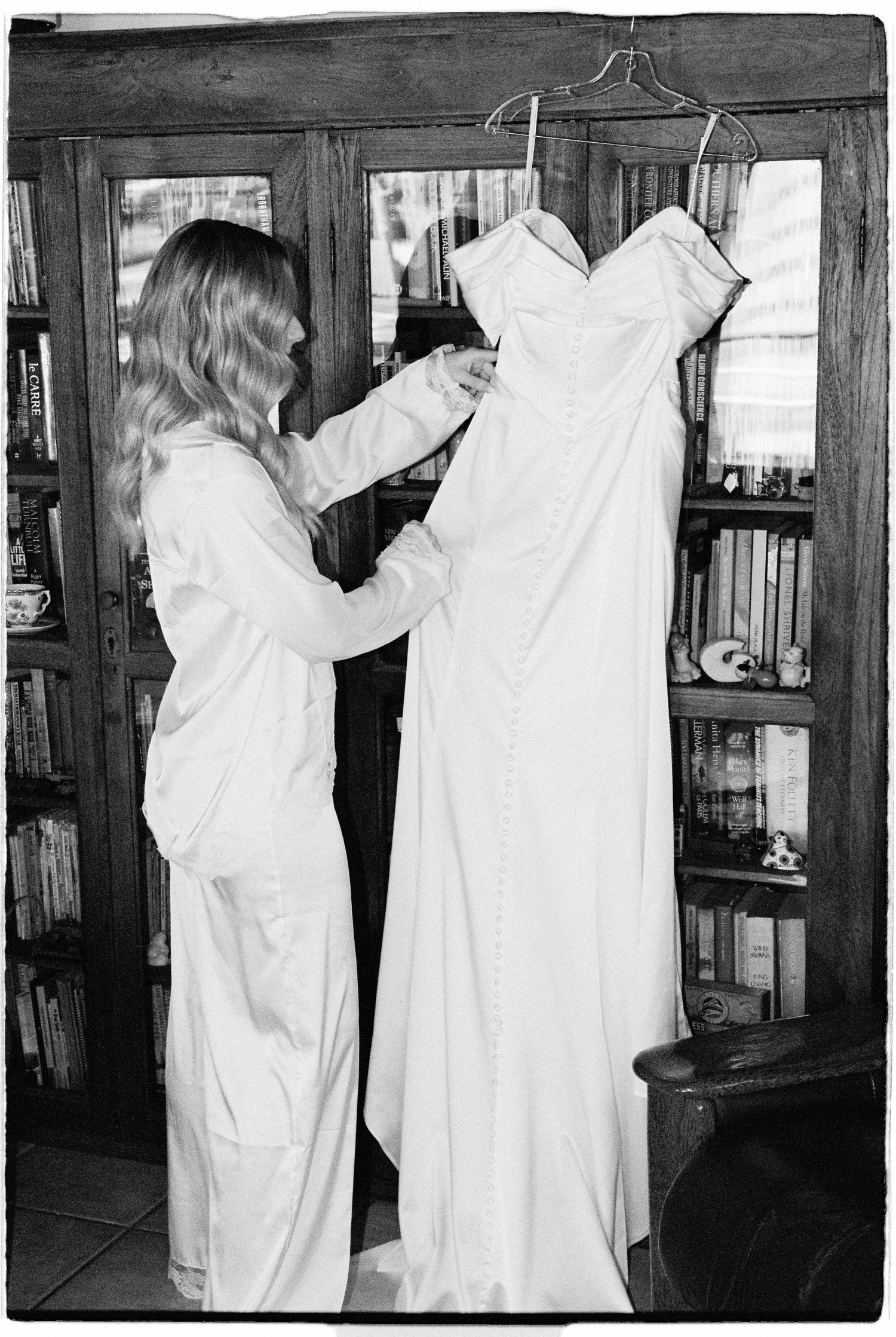 Black and white photo of blonde woman in white pajamas inspecting wedding dress on hanger by bookshelf.