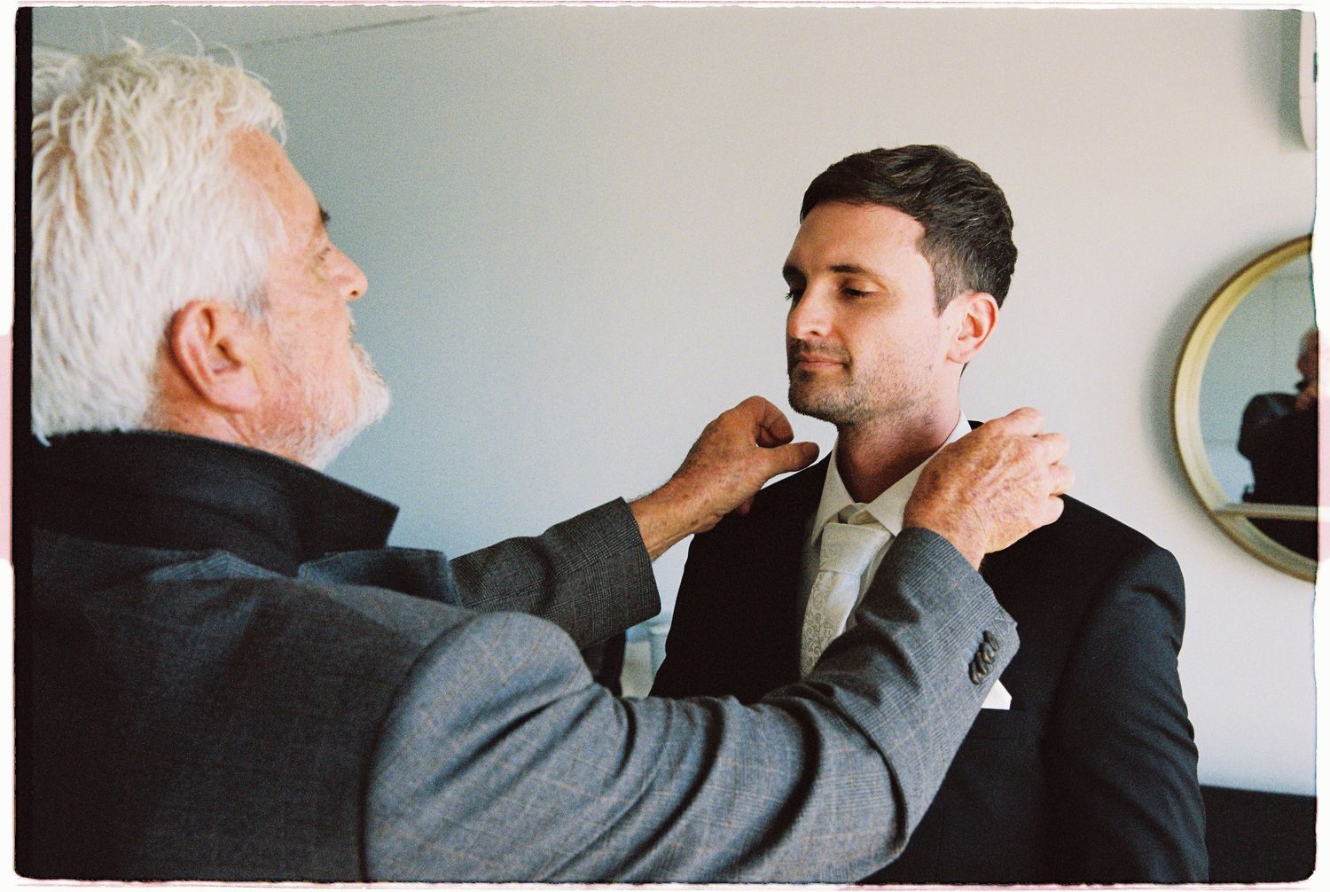 An older man adjusts the collar of a younger man in a dark suit, likely a father helping his son prepare for a wedding.