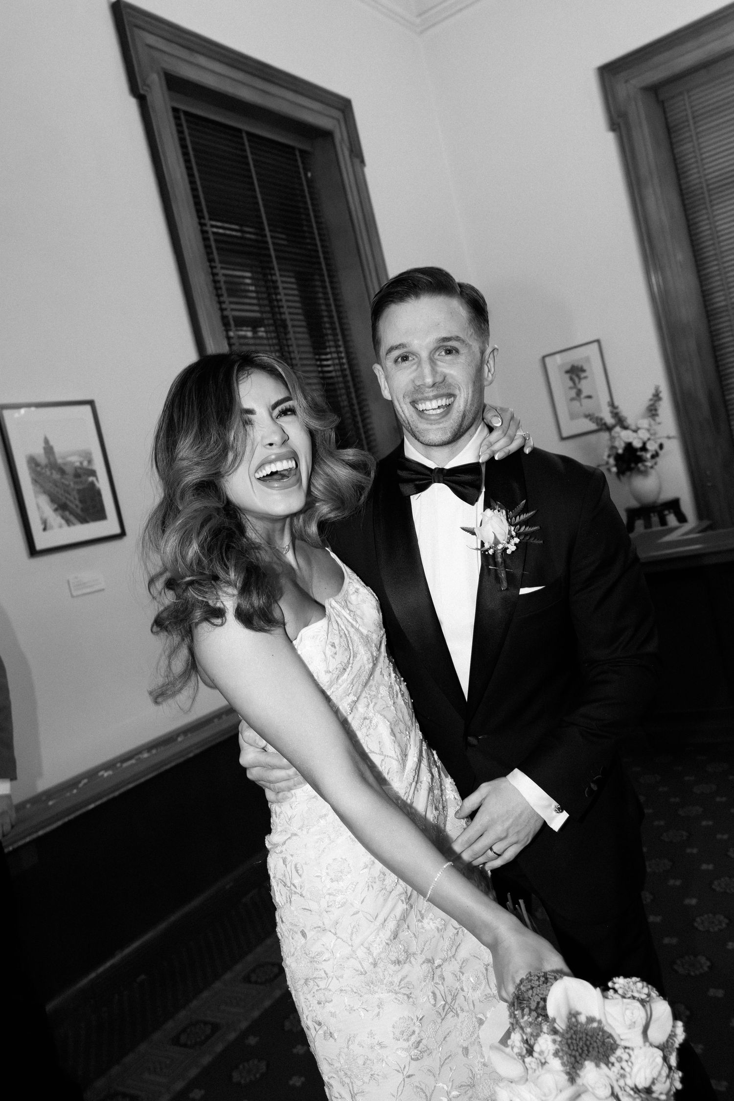 Black and white photo of a laughing bride in lace gown and groom in tuxedo embracing at their wedding.