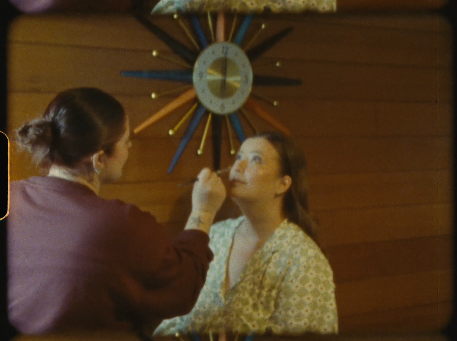 Woman applying makeup to another woman's face near a retro sunburst clock on a wood-paneled wall.