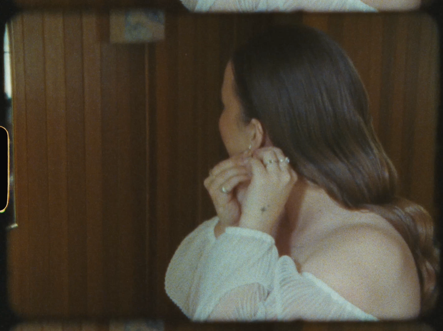 A woman with long dark hair touches her face while wearing a white top in a wood-paneled room.