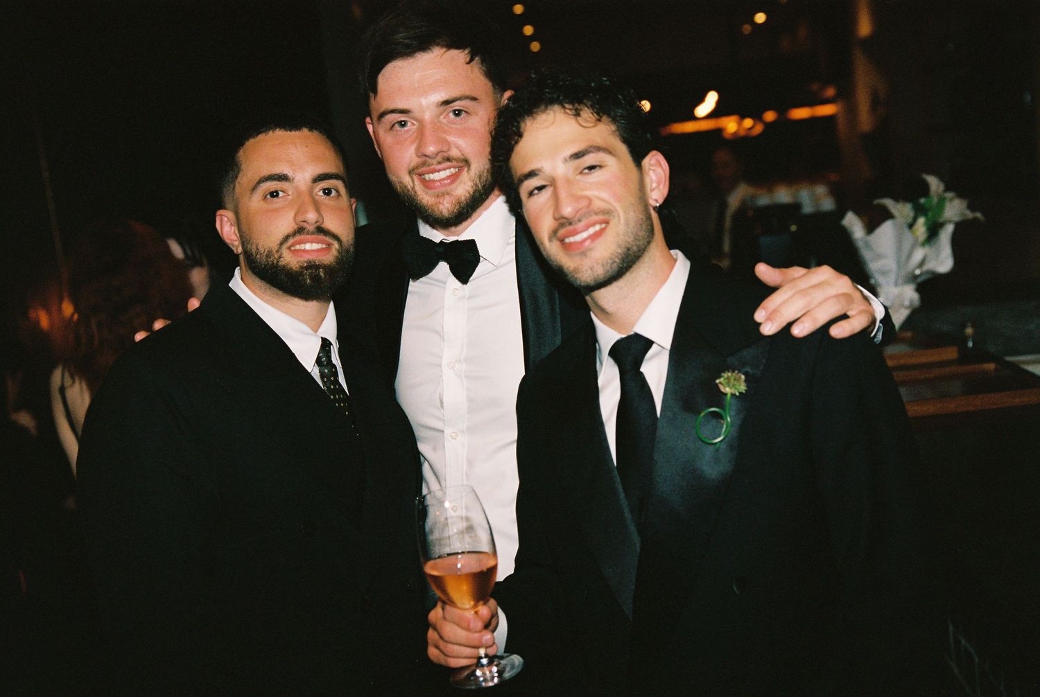 Three men in formal suits smiling together at a wedding reception event.