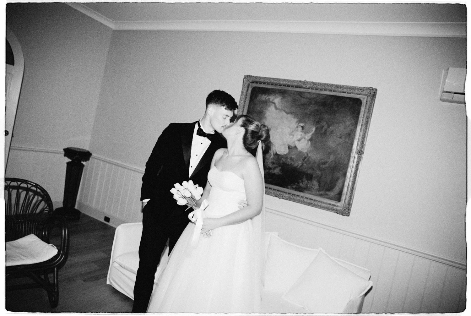 Black and white wedding photo of bride and groom sharing a kiss in an elegant room with a framed painting on the wall.