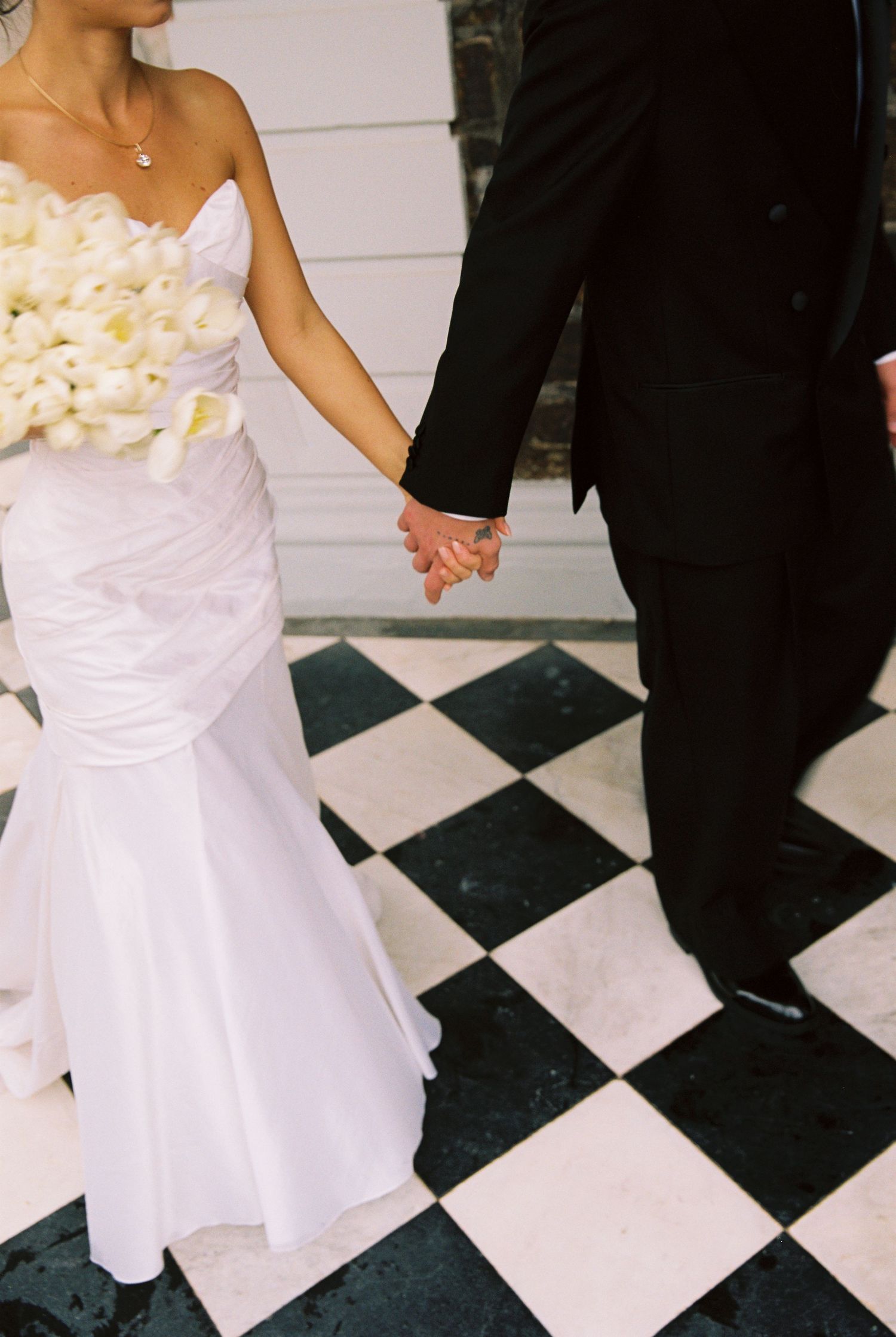 Bride and groom hold hands on black and white checkered floor, bride holds white bouquet.