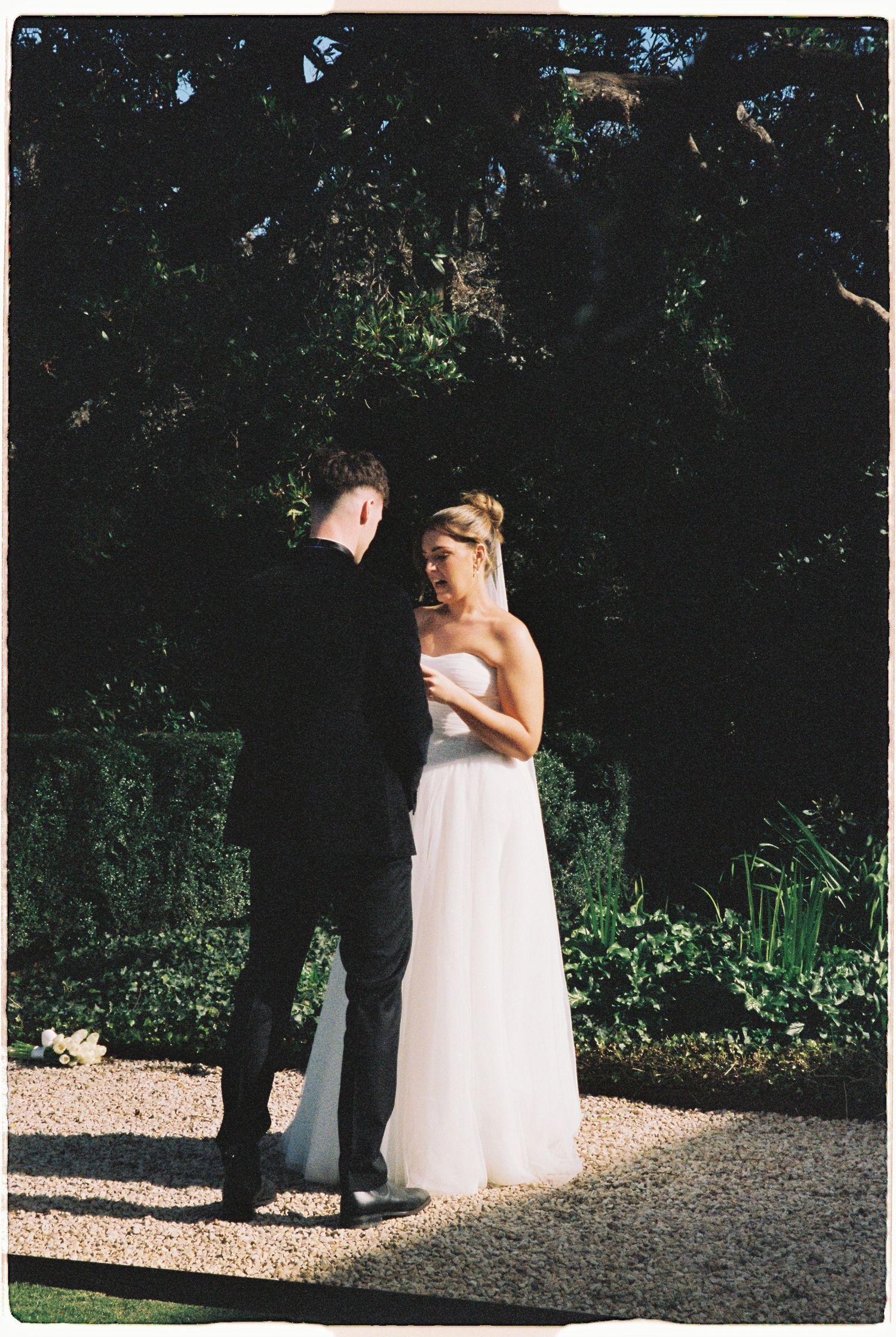 Bride in white strapless gown and groom in black suit share intimate moment in sunlit garden ceremony.