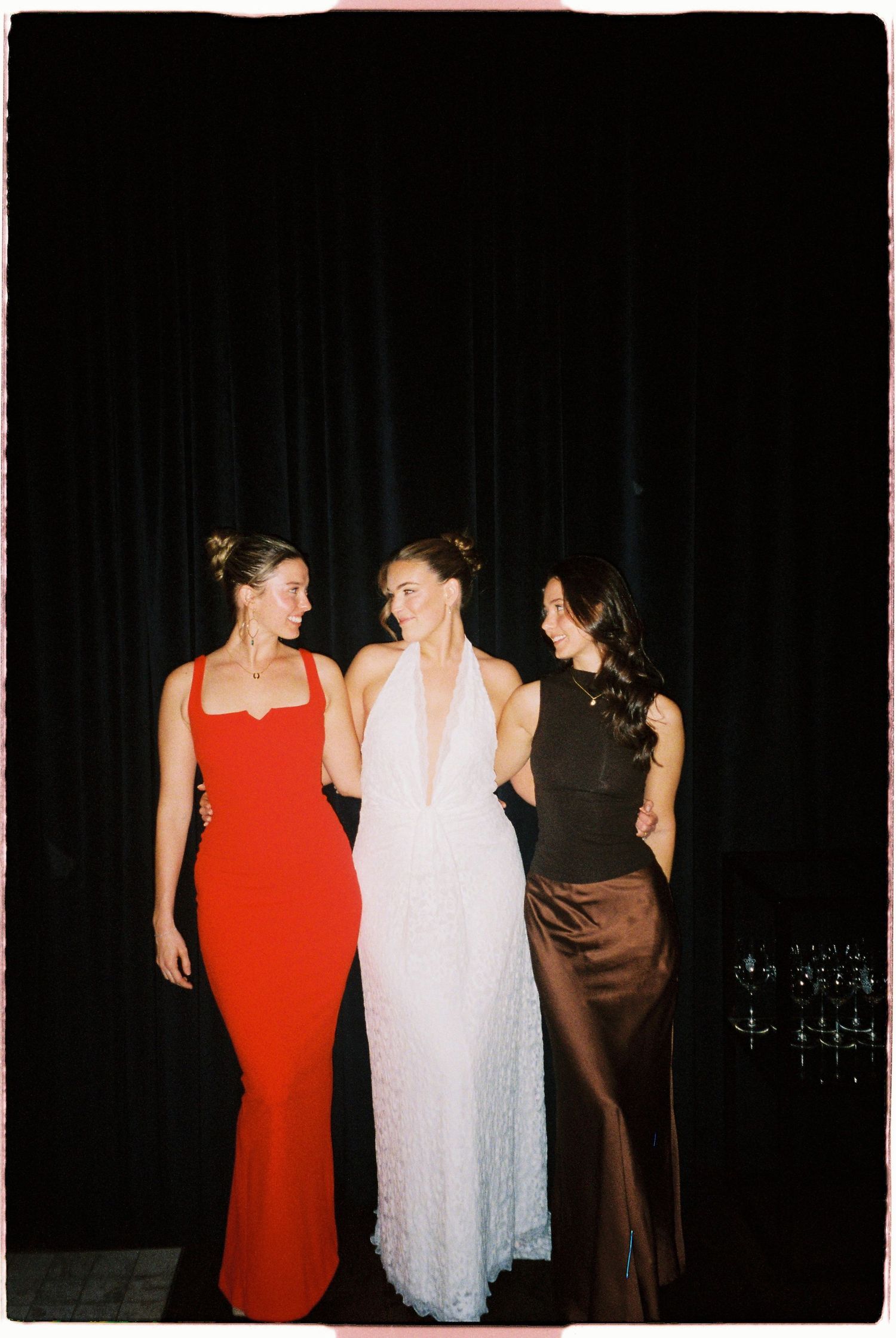 Three women in elegant gowns posing together in red, white, and brown dresses.