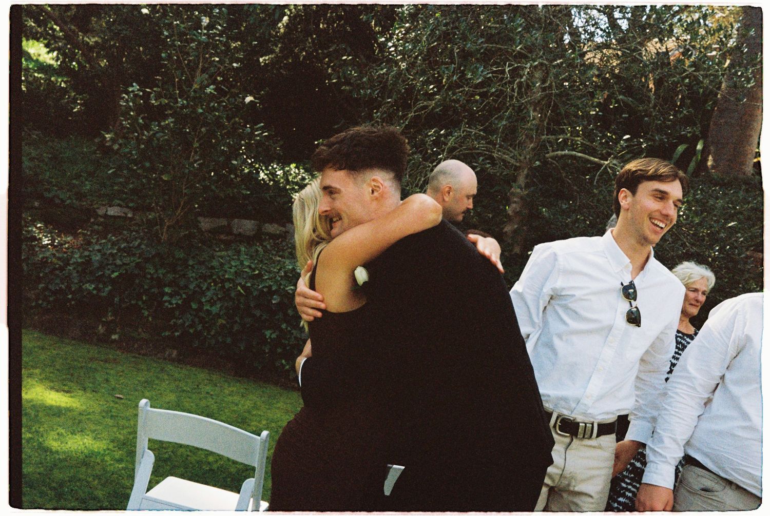 Groom embraces wedding guest warmly while another smiling guest looks on at outdoor reception.
