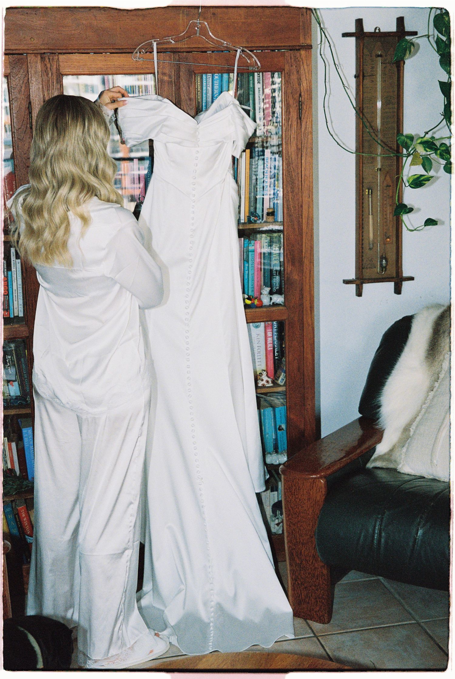 Blonde woman holding a white wedding dress on a hanger in front of a wooden bookshelf.
