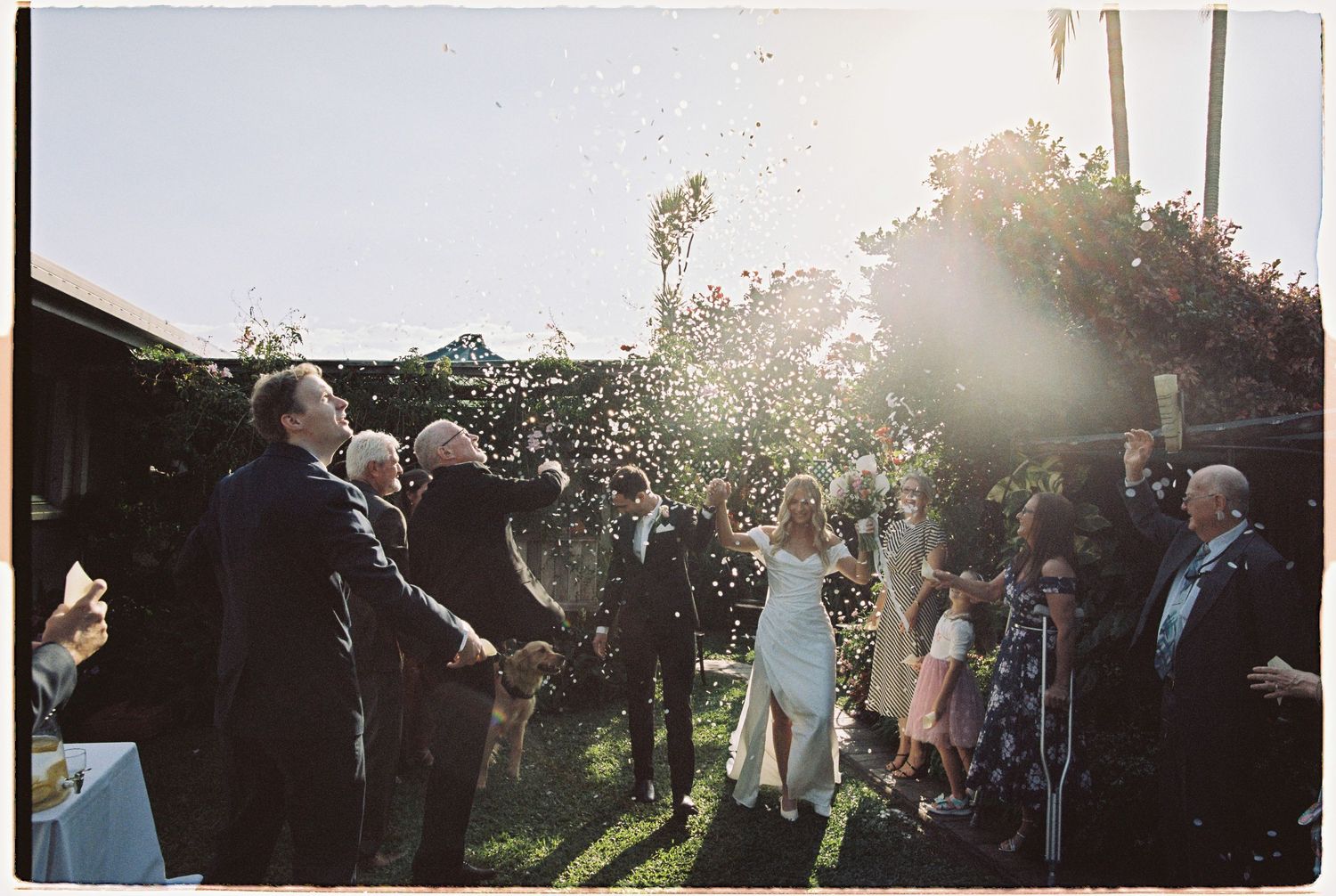 Wedding guests toss confetti as bride and groom exit ceremony in sunny backyard garden celebration.