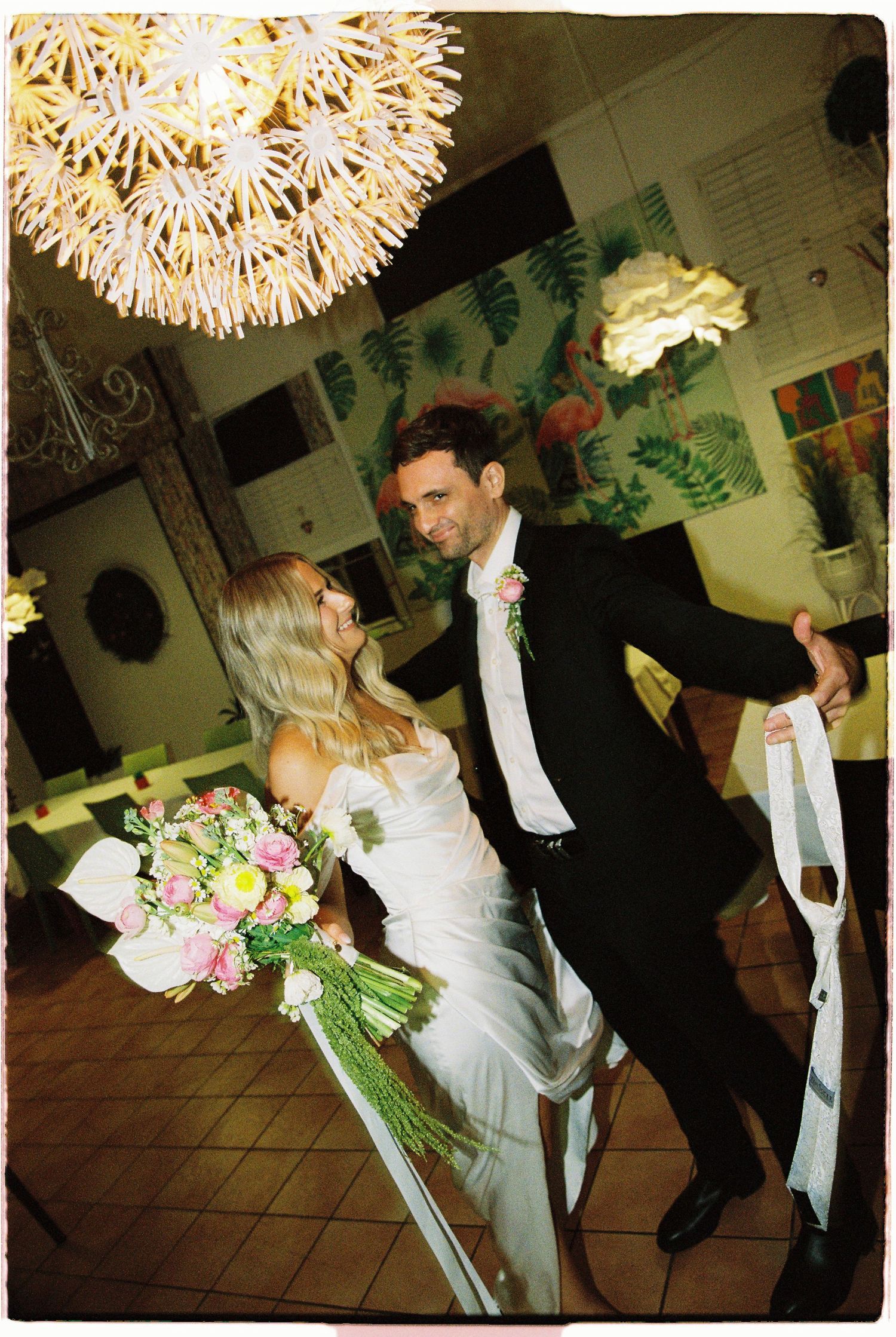 Bride and groom dancing indoors under decorative chandelier, bride holding pink and white floral bouquet.