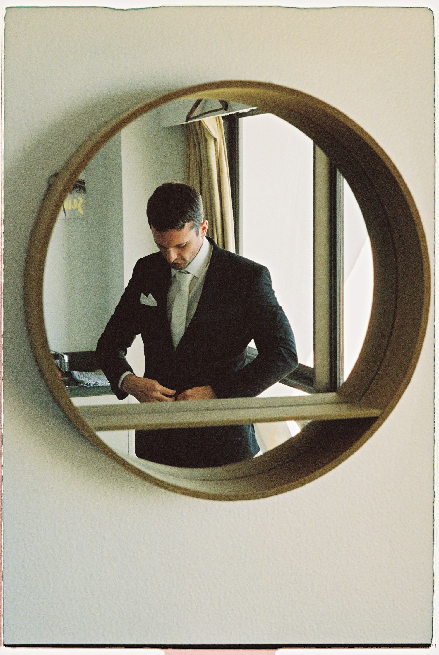 Groom in dark suit adjusting attire reflected in circular gold mirror in hotel room.