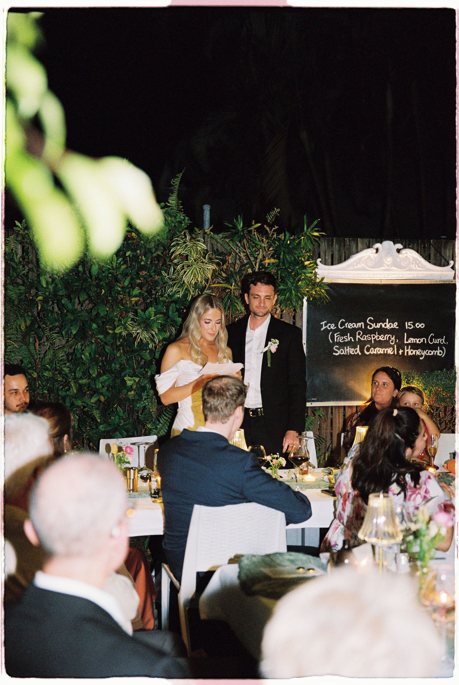 Bride and groom stand at wedding reception dinner table giving speech, guests seated around floral arrangements at night.