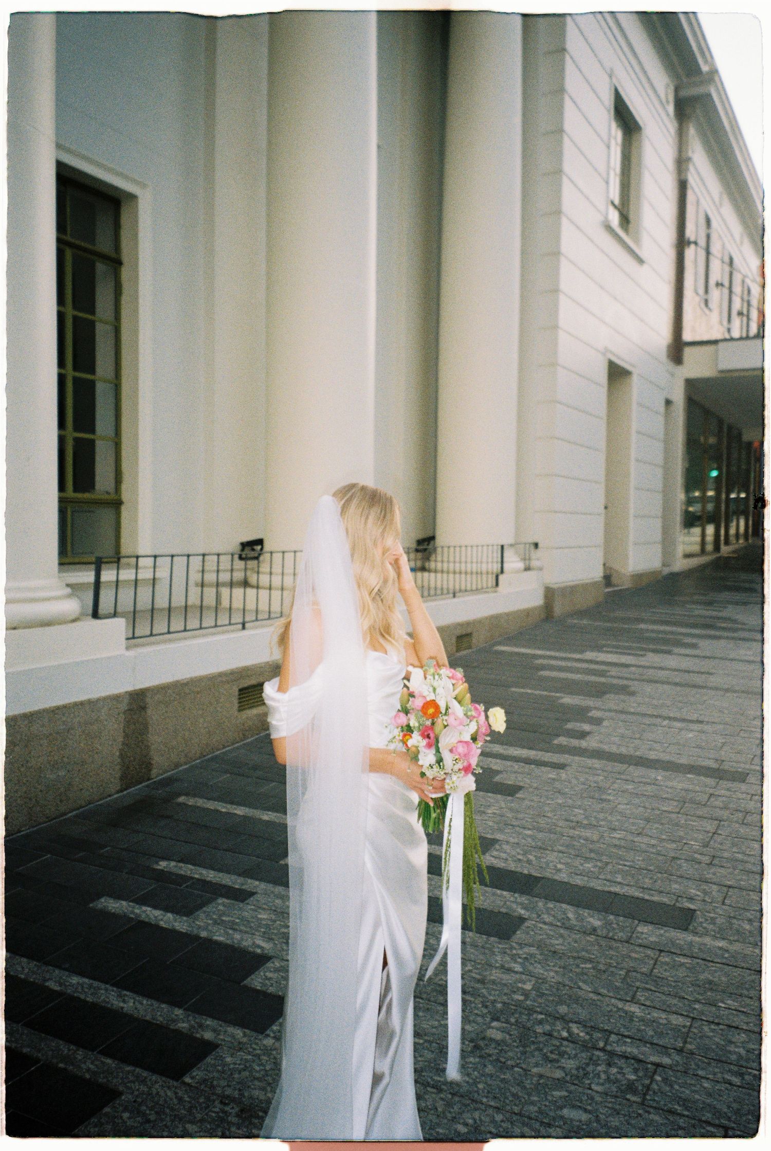 Bride in white gown and veil walking along a classical building exterior holding a floral bouquet.