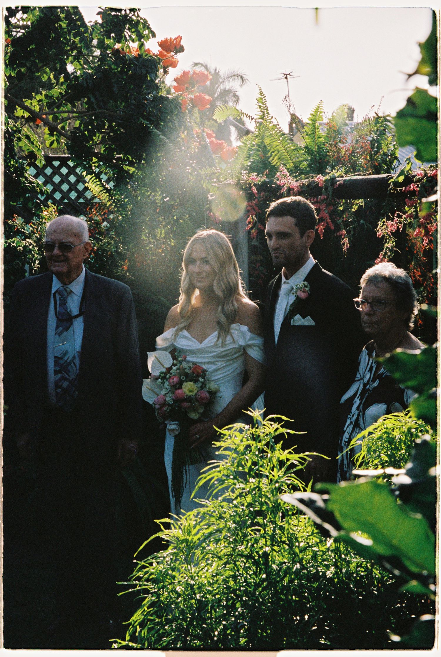 Bride holding a colorful bouquet stands with groom and elderly guests in a sunlit garden wedding.