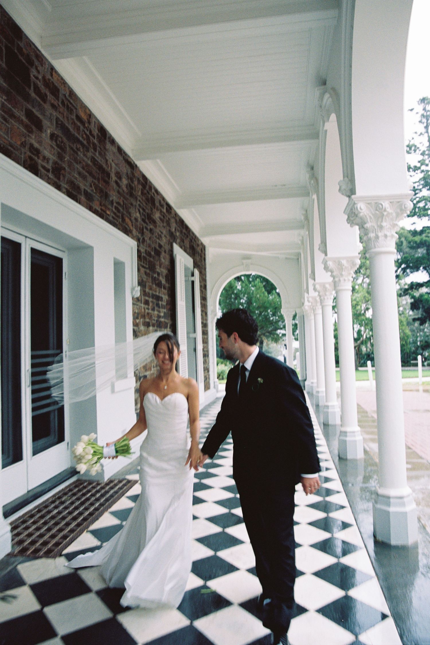 Bride and groom walking on black and white checkered floor of elegant venue colonnade.