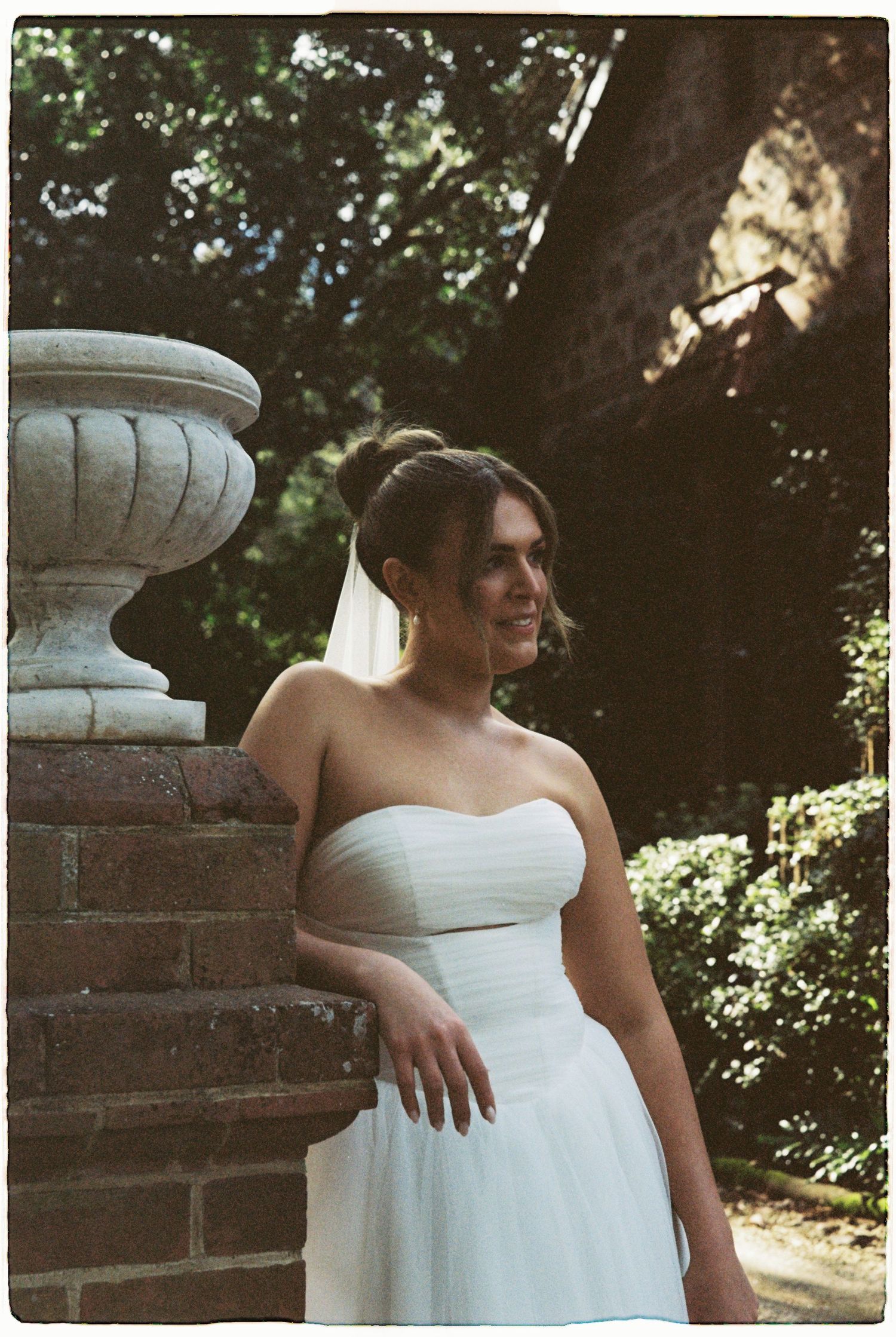 Beautiful bride in strapless white gown poses beside ornate stone urn and brick wall outdoors.