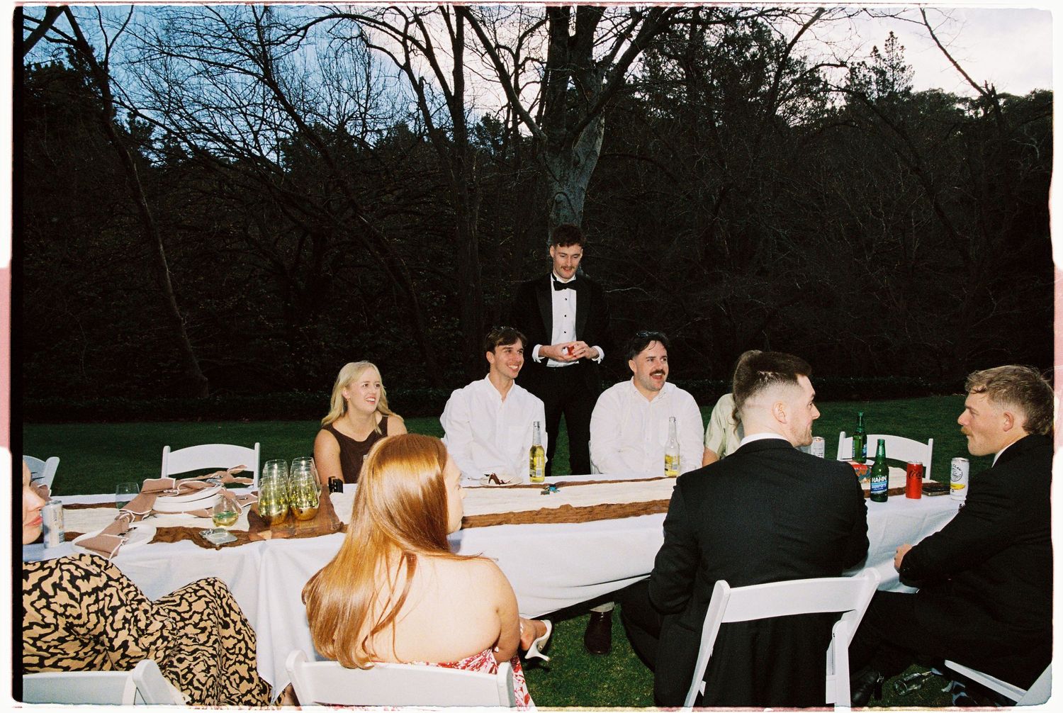 Wedding guests dining at an outdoor evening reception table while a server presents a dish.