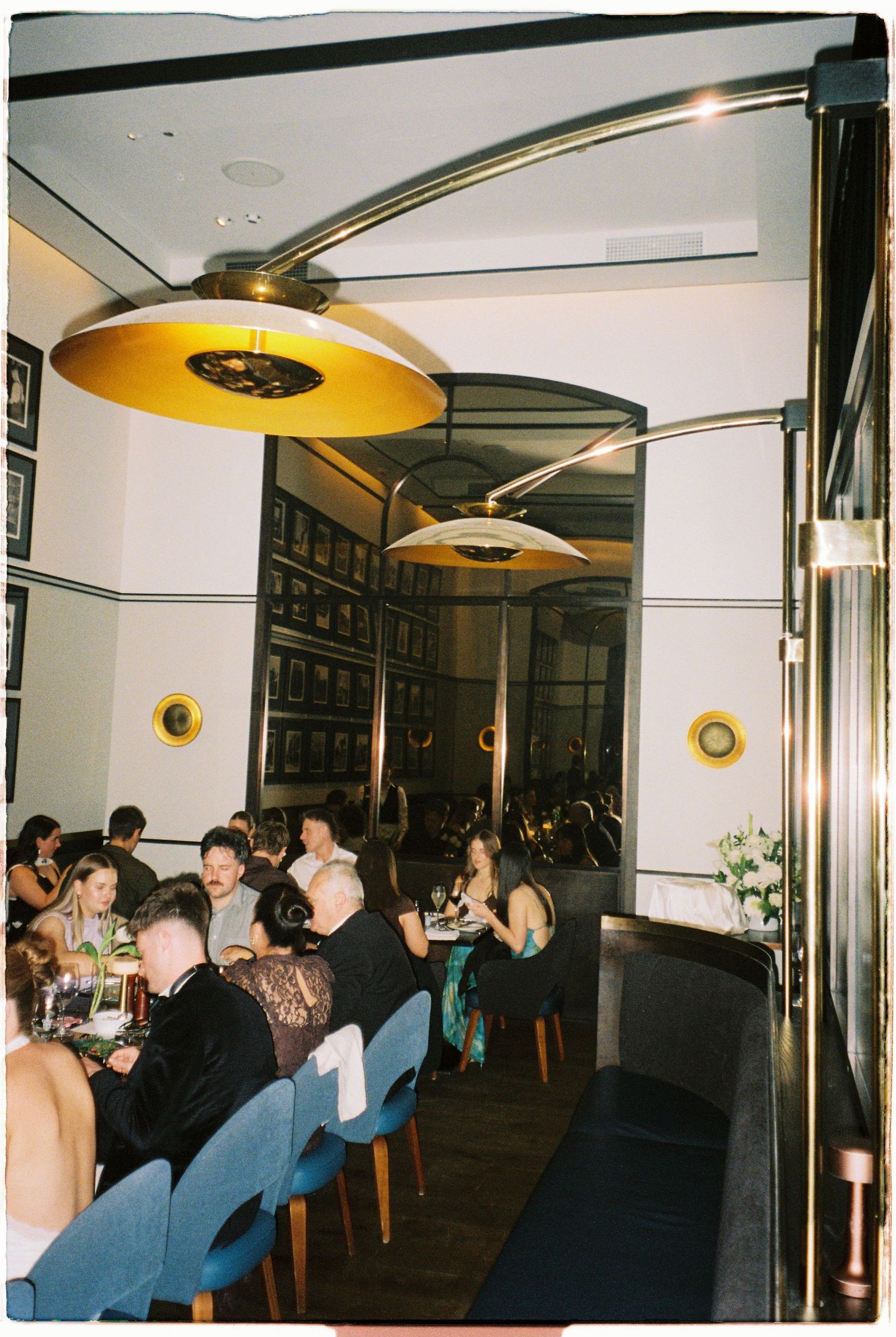 Wedding reception dinner guests seated in a stylish restaurant with gold pendant lighting.