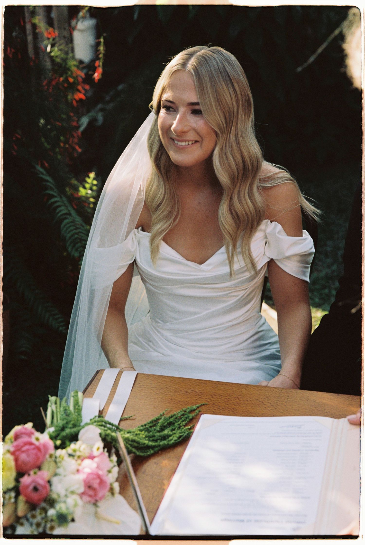 Smiling blonde bride in off-shoulder white gown with veil signing wedding register outdoors.