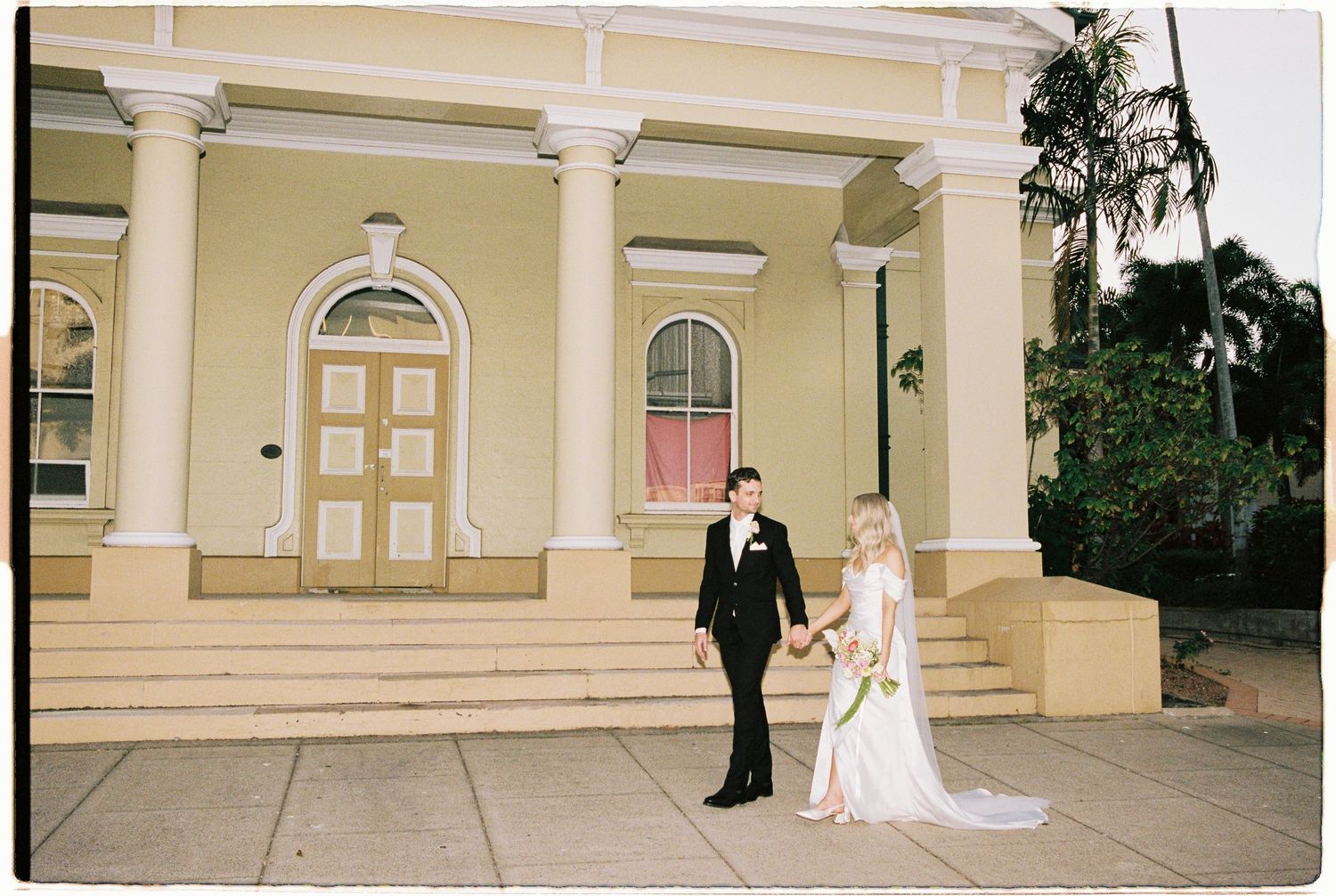 Bride and groom holding hands in front of a grand yellow colonial building with white columns.