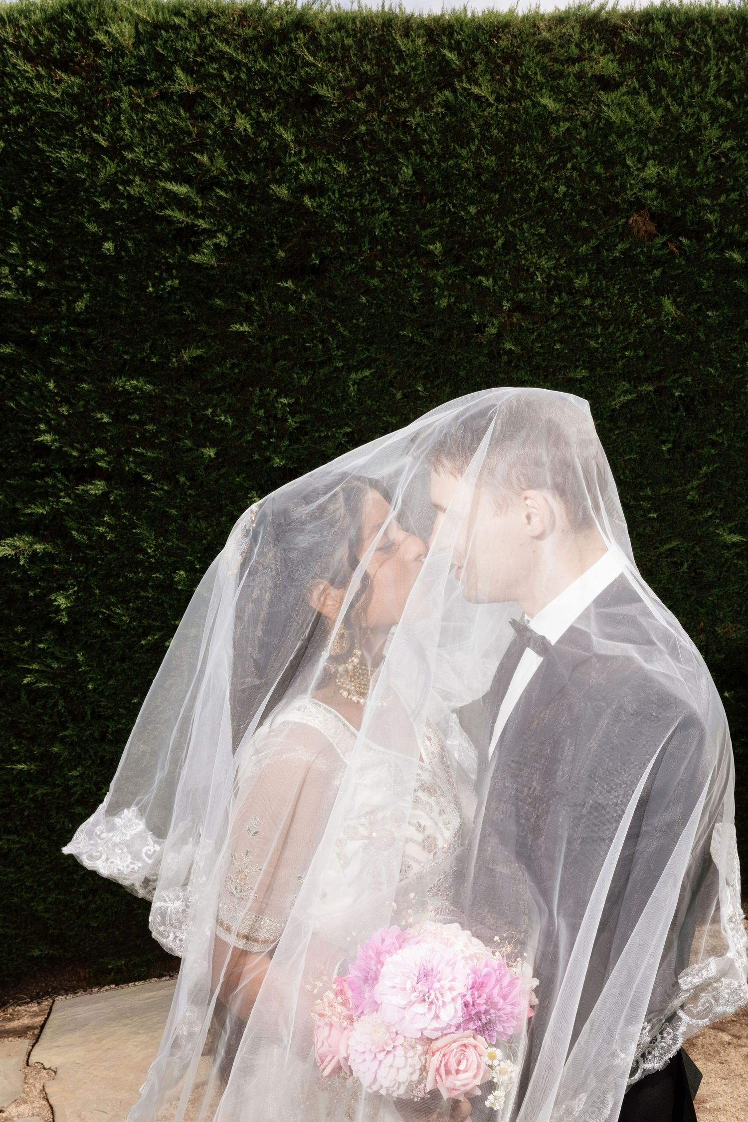 Bride and groom sharing romantic moment hidden beneath elegant lace wedding veil, bride holding pink bouquet.