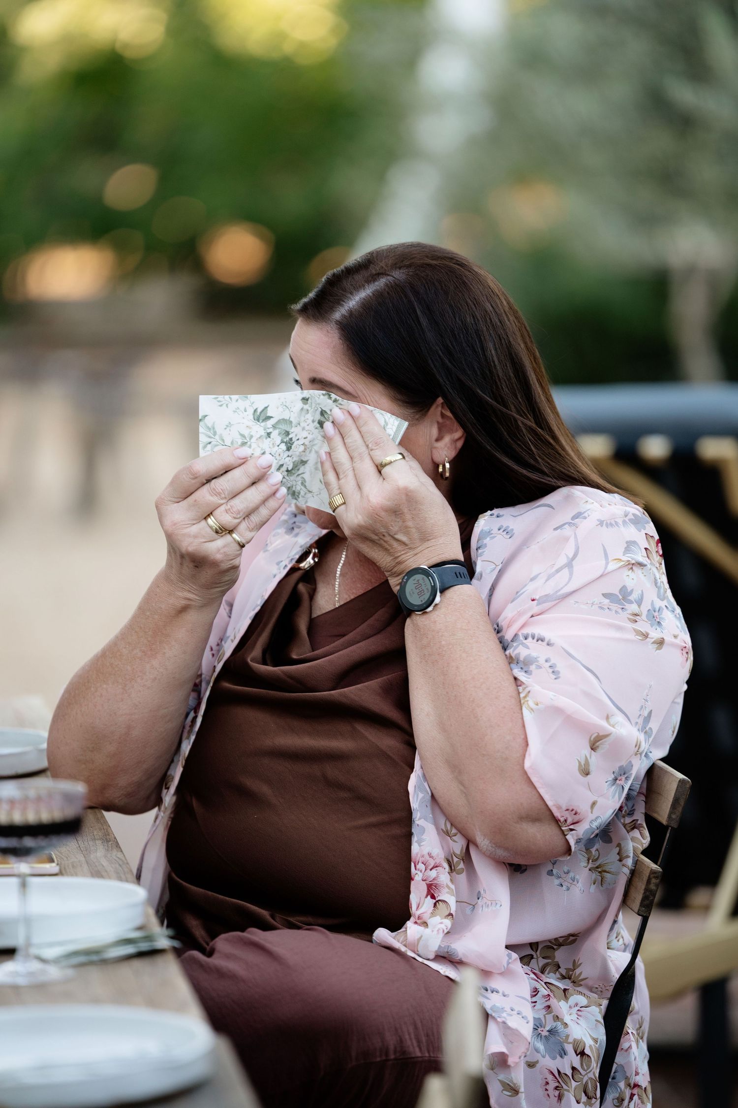Emotional wedding guest dabbing tears with floral napkin at outdoor reception table.
