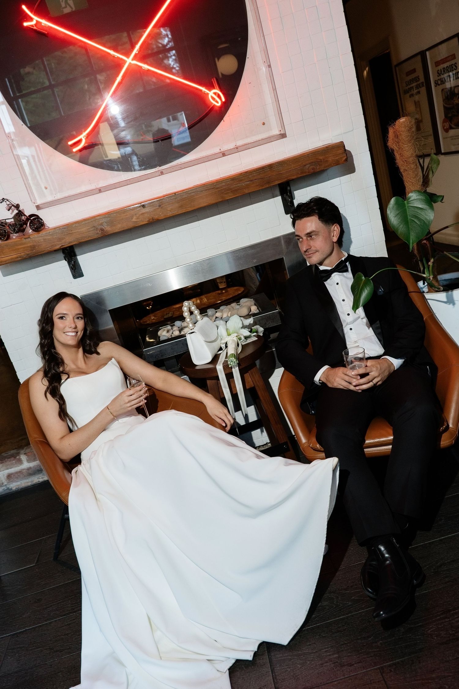 Bride in white strapless gown and groom in black tuxedo sitting in leather chairs by a fireplace.