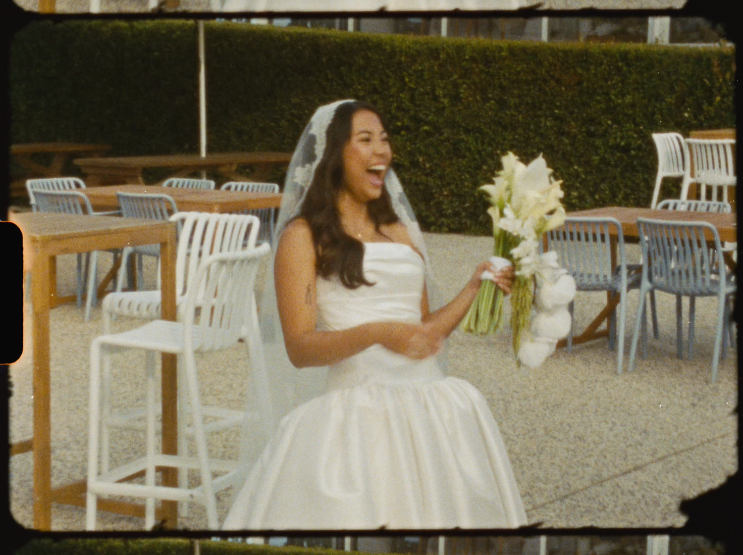 Excited bride in white strapless gown holding bouquet and laughing in outdoor venue with chairs.