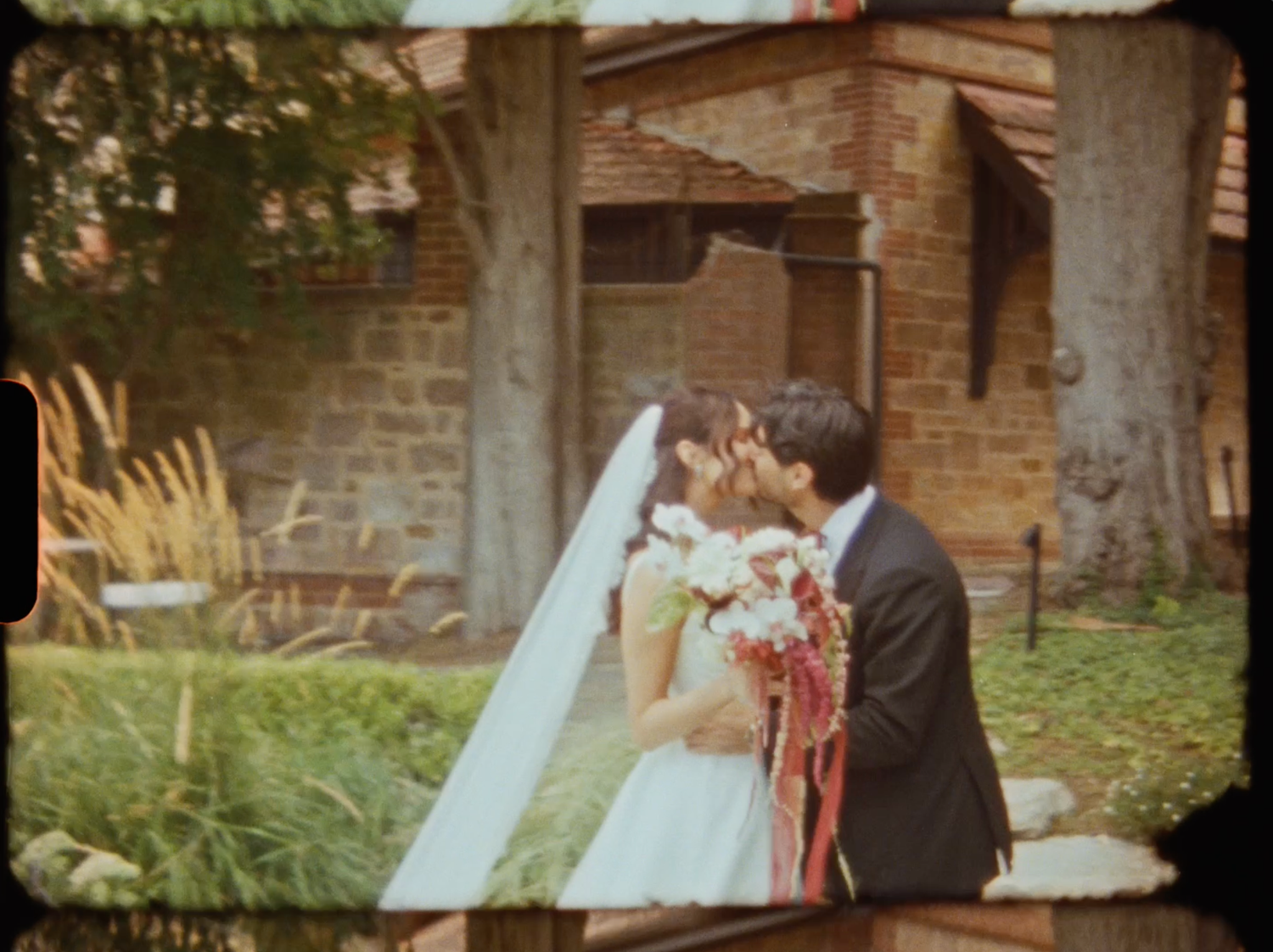 Bride and groom sharing a kiss surrounded by wedding bouquet with red ribbons outdoors.