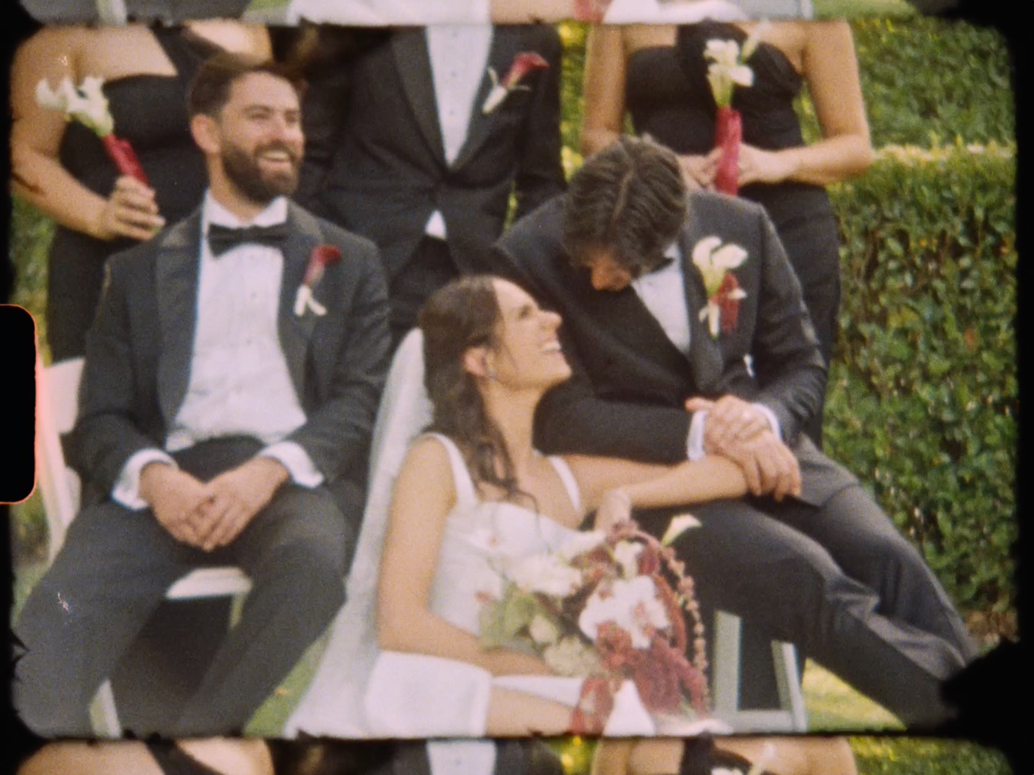 Groom lovingly leans toward smiling bride holding bouquet surrounded by laughing wedding party outdoors.