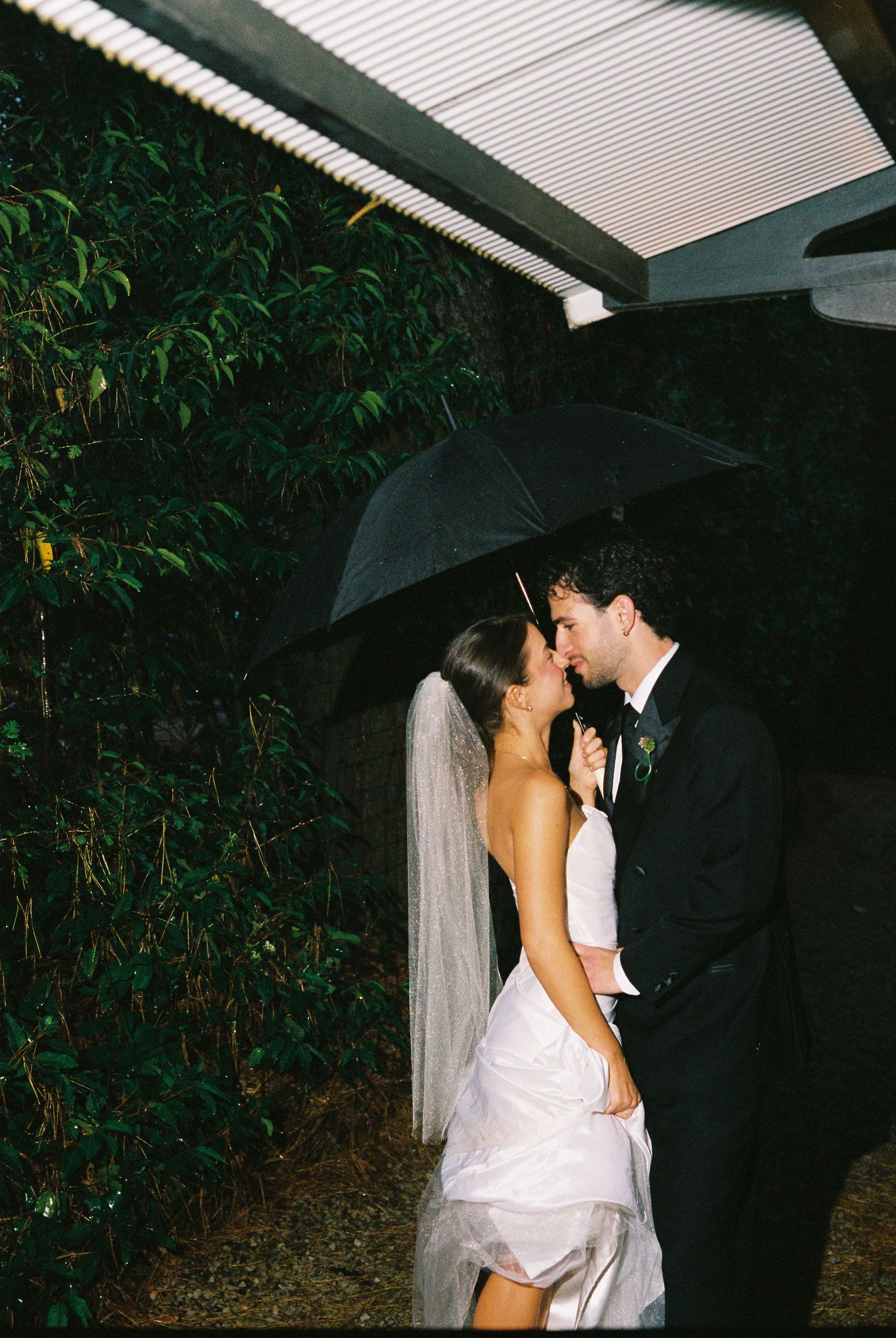 Bride and groom sharing a romantic kiss under a black umbrella in the rain.