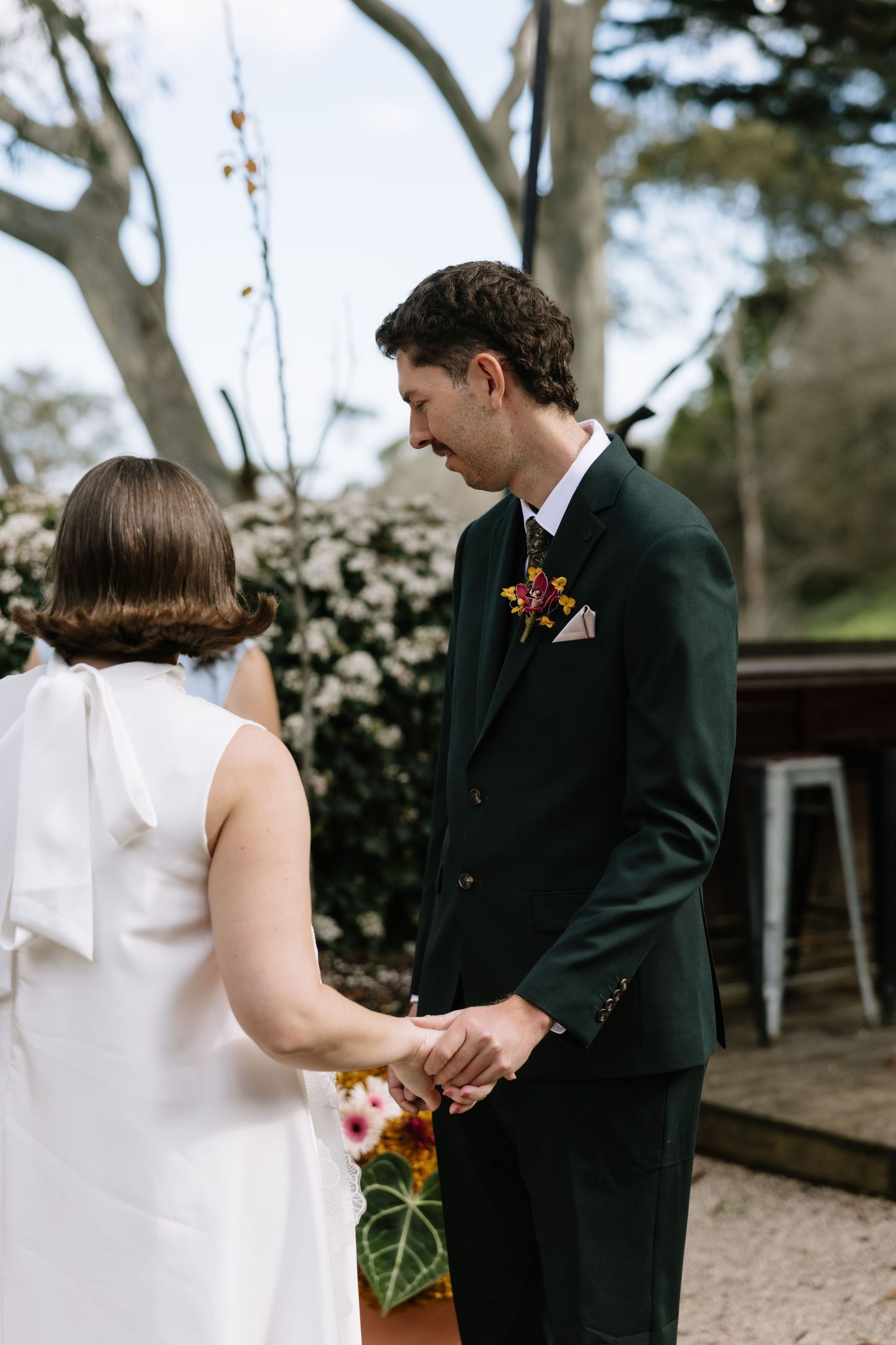 Groom in dark green suit holds bride's hands during outdoor wedding ceremony with floral backdrop.