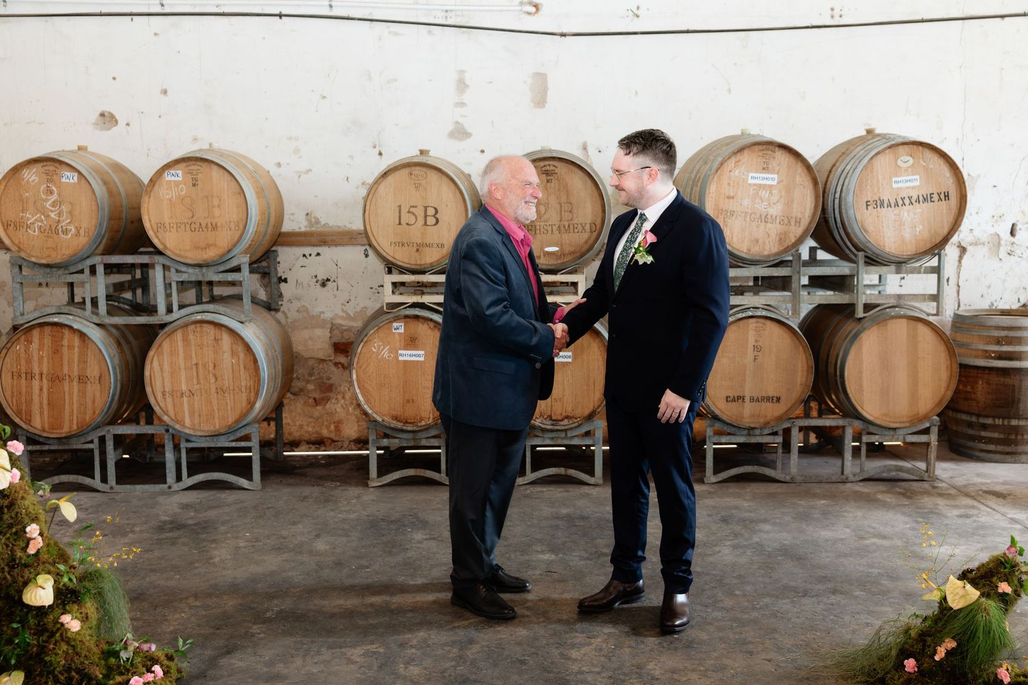 Two men in suits shaking hands in front of wooden wine barrels at a winery wedding.