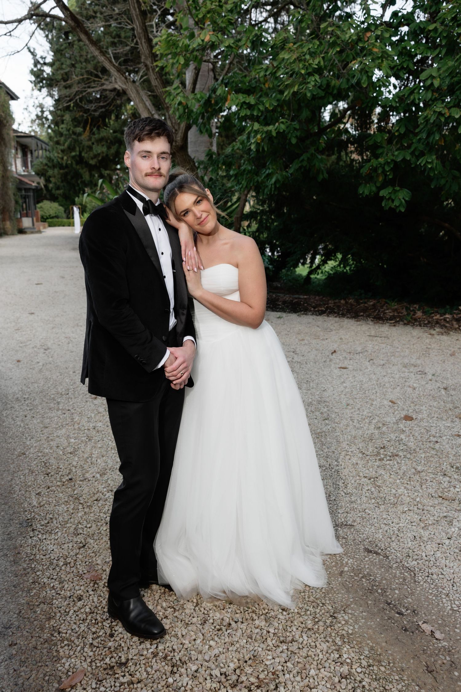 Bride in strapless white tulle gown leans on groom in black tuxedo outdoors on gravel path.