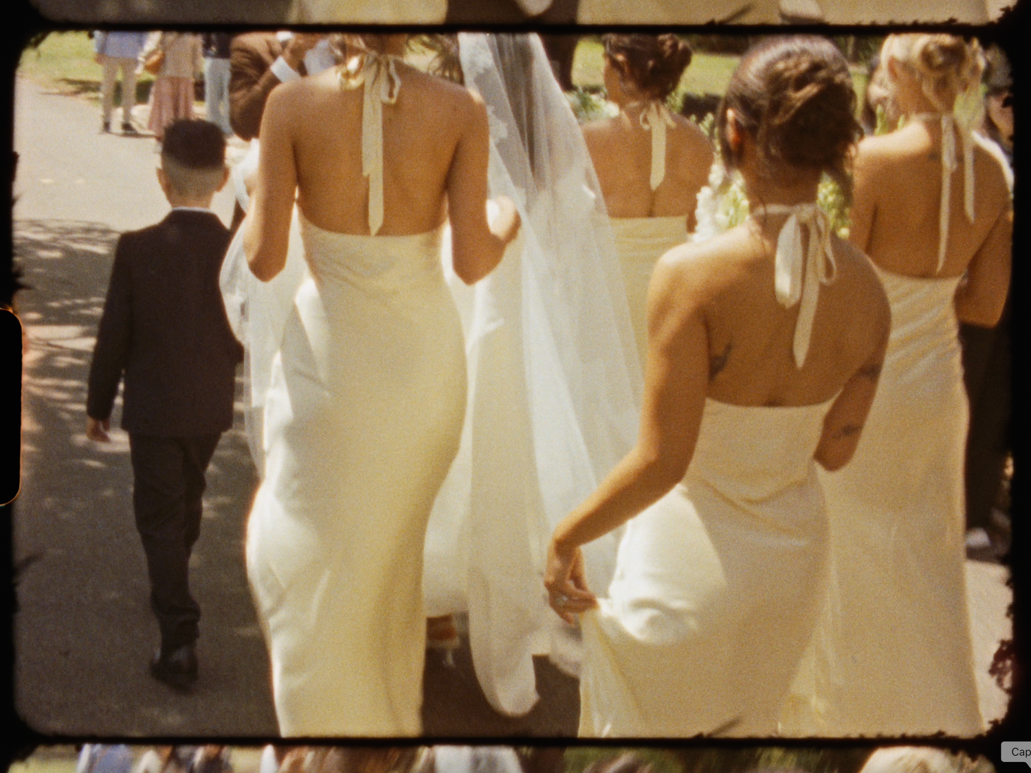 Bride and bridesmaids in white halter gowns walk with a young boy in a suit at an outdoor wedding ceremony.
