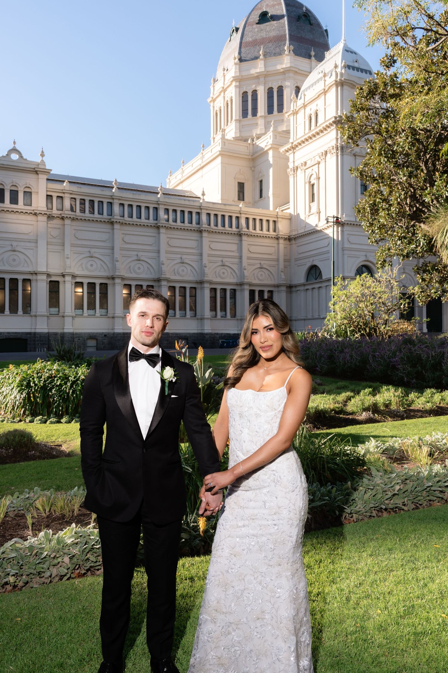 Bride and groom posing in formal wedding attire in front of a grand historic building with manicured gardens.