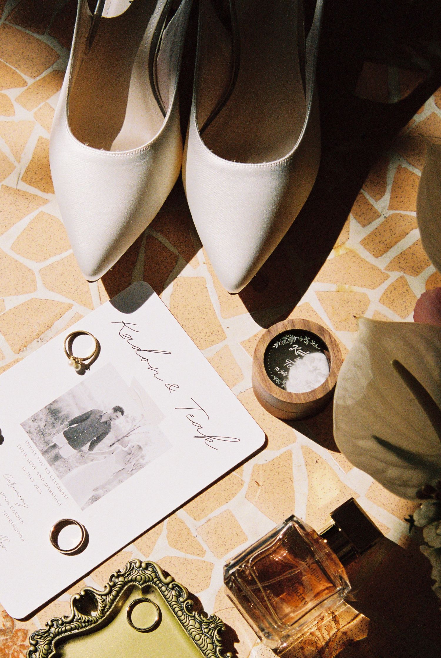 Flat lay of white bridal heels, wedding invitation, gold rings, perfume bottle, and wooden dish on a tiled floor.